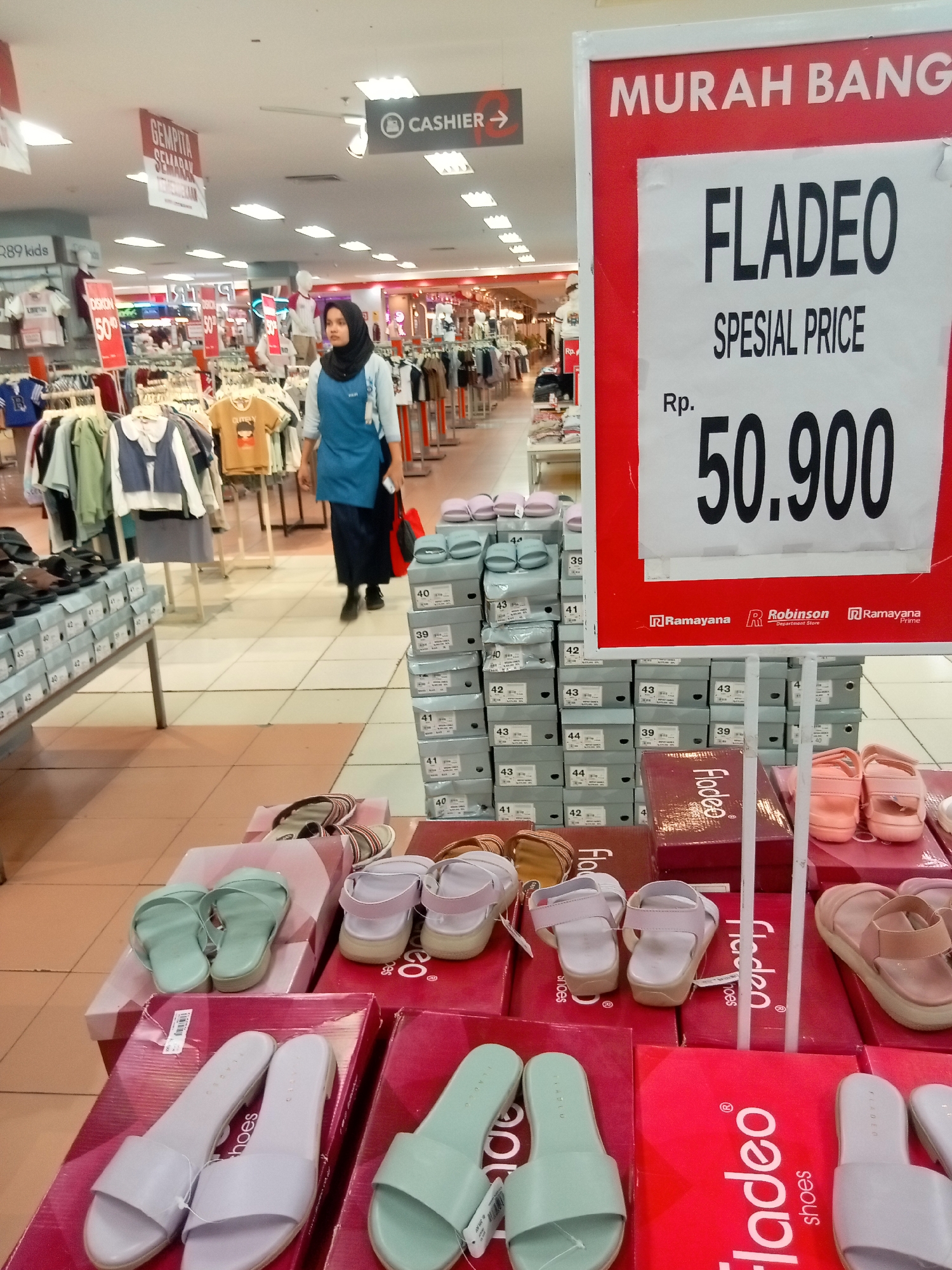 This image shows a retail store, likely a department store or shoe store, in Batam, Indonesia, based on the language and brands visible. In the foreground, there is a display of various footwear, including sandals and flip-flops, arranged on top of boxes. The footwear is predominantly in pastel colors like mint green, light purple, and beige. The boxes are red and white and some have branding like "Fladeo" and "Ramayana". Behind the shoe display, there are stacks of shoeboxes, many labeled with sizes like 39, 40, 41, 42, and 43.
In the middle ground, a woman wearing a dark hijab and a light blue uniform shirt is walking through the store, carrying a red bag. She appears to be an employee. Further in the background, clothing racks with various garments are visible, along with signage that includes the word "CASHIER". The store appears well-lit with fluorescent ceiling lights. A large red and white promotional sign is prominently displayed on the right side of the image, advertising "MURAH BANGET" (very cheap) and "FLADEO SPESIAL PRICE RP. 50.900". The flooring is tiled.
