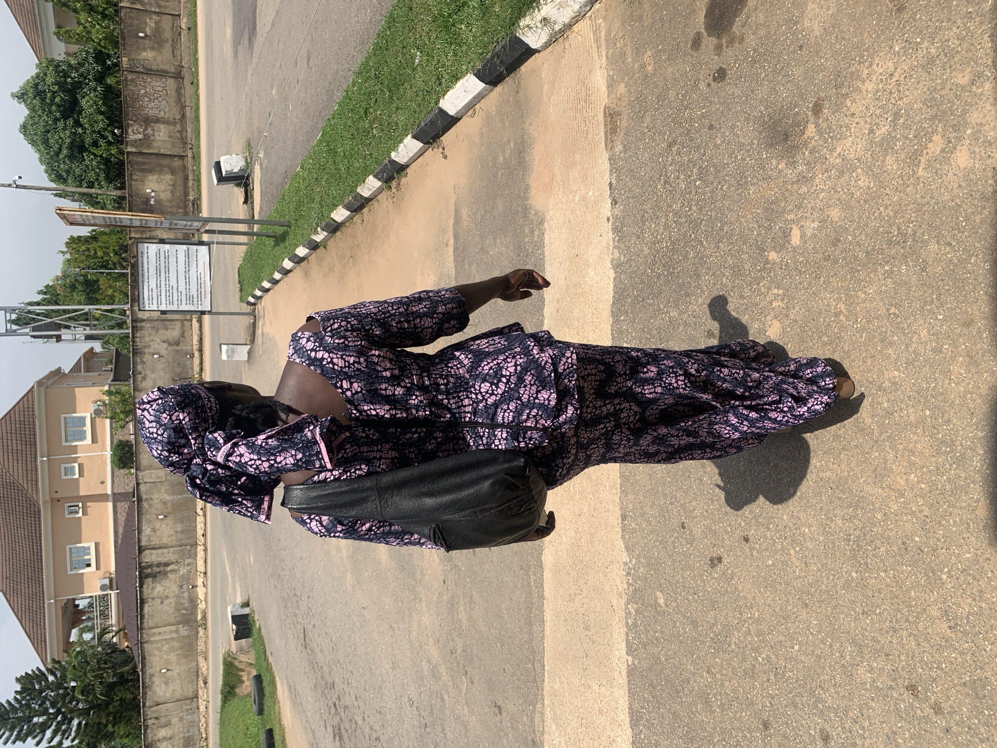 A full-length shot from behind shows a woman walking on a paved sidewalk in Abuja, Nigeria. She is wearing a pink and navy blue patterned dress and matching headscarf, with a black leather shoulder bag slung over her left shoulder. The sidewalk is bordered by a grassy area with a black and white striped curb. In the background, a large, tan house with a brown roof is visible behind a concrete wall. Trees and a street sign are also present in the scene. The sky is overcast.