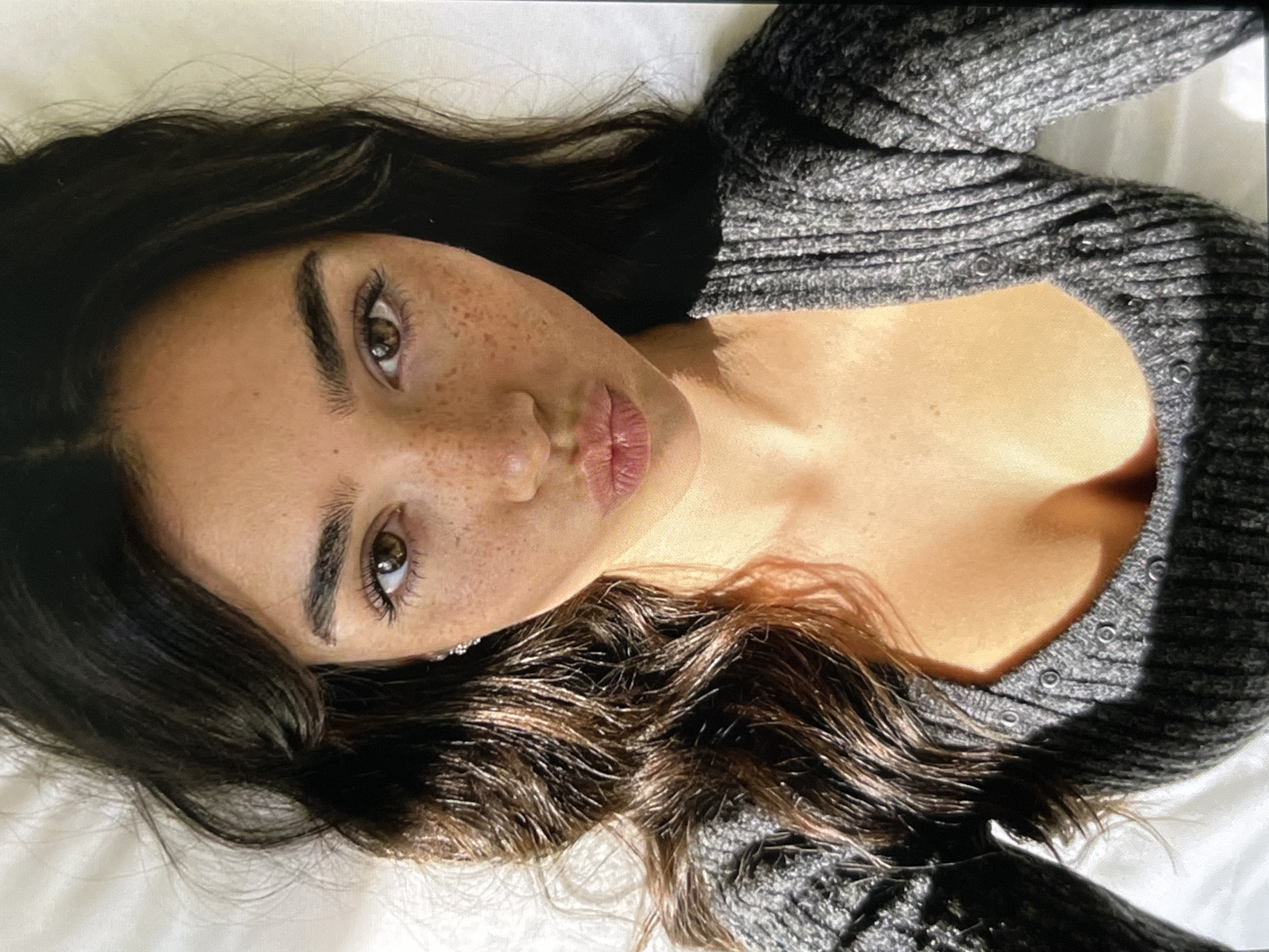 A close-up, overhead shot shows a young woman with dark brown hair and fair skin with freckles. She is wearing a grey, ribbed sweater and has her lips pursed. The lighting is warm and casts shadows, suggesting natural light. The background is a plain, white surface, possibly a bed or couch.