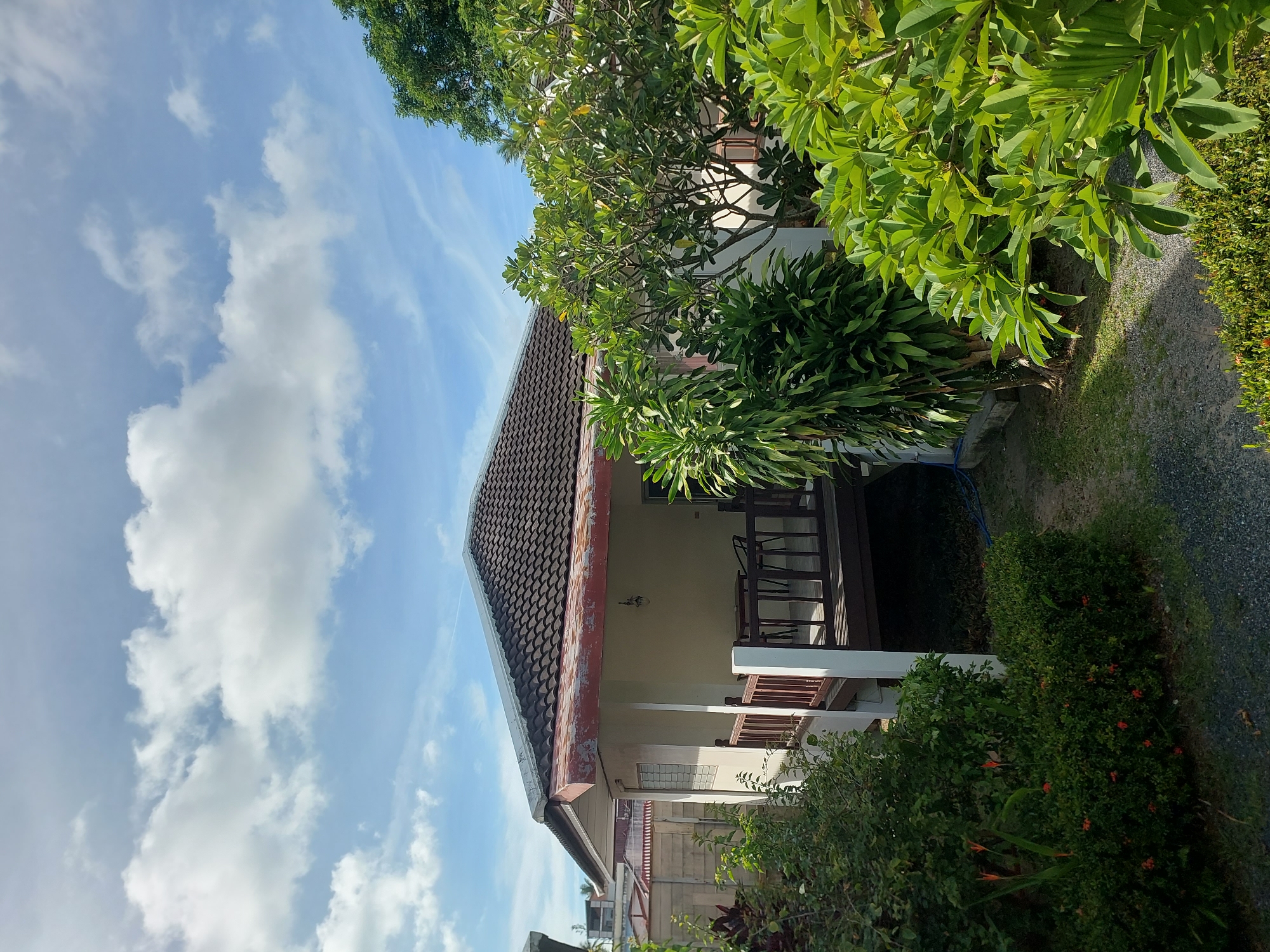 A one-story house with a tiled roof is visible in Baan Mae Nam, Thailand, under a partly cloudy sky. The house has a cream-colored exterior, a covered porch with a wooden railing, and a small outdoor seating area. Lush green foliage, including trees and bushes, surrounds the house. A gravel path leads to the porch. The scene appears to be peaceful and idyllic.