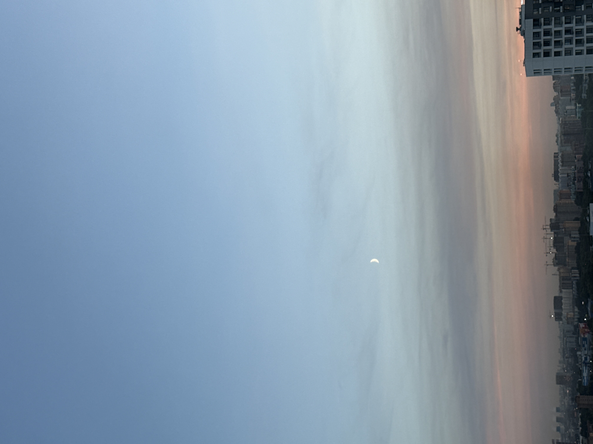The image depicts a twilight view over the city of Lyubertsy, Russia. The sky is a gradient of pale blue to soft pink and orange hues, with a thin crescent moon visible in the upper portion of the frame. Wispy clouds are scattered across the sky, particularly in the mid-ground where they are tinged with the colors of sunset. The foreground is dominated by the cityscape, showing a dense collection of buildings. In the far distance, there are some industrial structures with visible smokestacks. Numerous construction cranes are scattered throughout the city, indicating ongoing development. A tall, modern white building with many windows stands prominently in the right foreground. The overall scene is one of an urban environment at dusk.