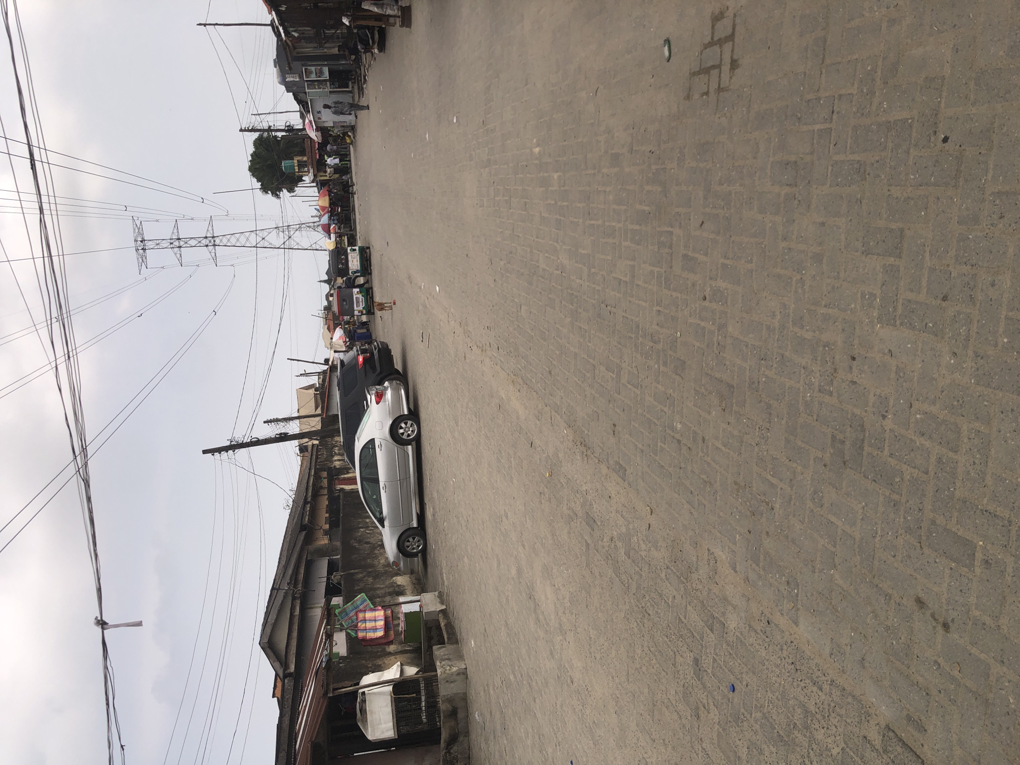 The image depicts a street scene in Ado, Nigeria, characterized by a paved road and buildings on either side. In the foreground, the road is paved with interlocking blocks, showing signs of wear and tear. The midground reveals several vehicles, including a silver sedan parked on the left and a black SUV. In the distance, a bustling market area is visible with stalls, umbrellas, and a crowd of people. A large electricity pylon stands prominently in the background, with numerous overhead wires crisscrossing the sky. The sky is overcast, suggesting a cloudy day. The overall environment appears to be a busy urban area with a mix of residential and commercial structures.