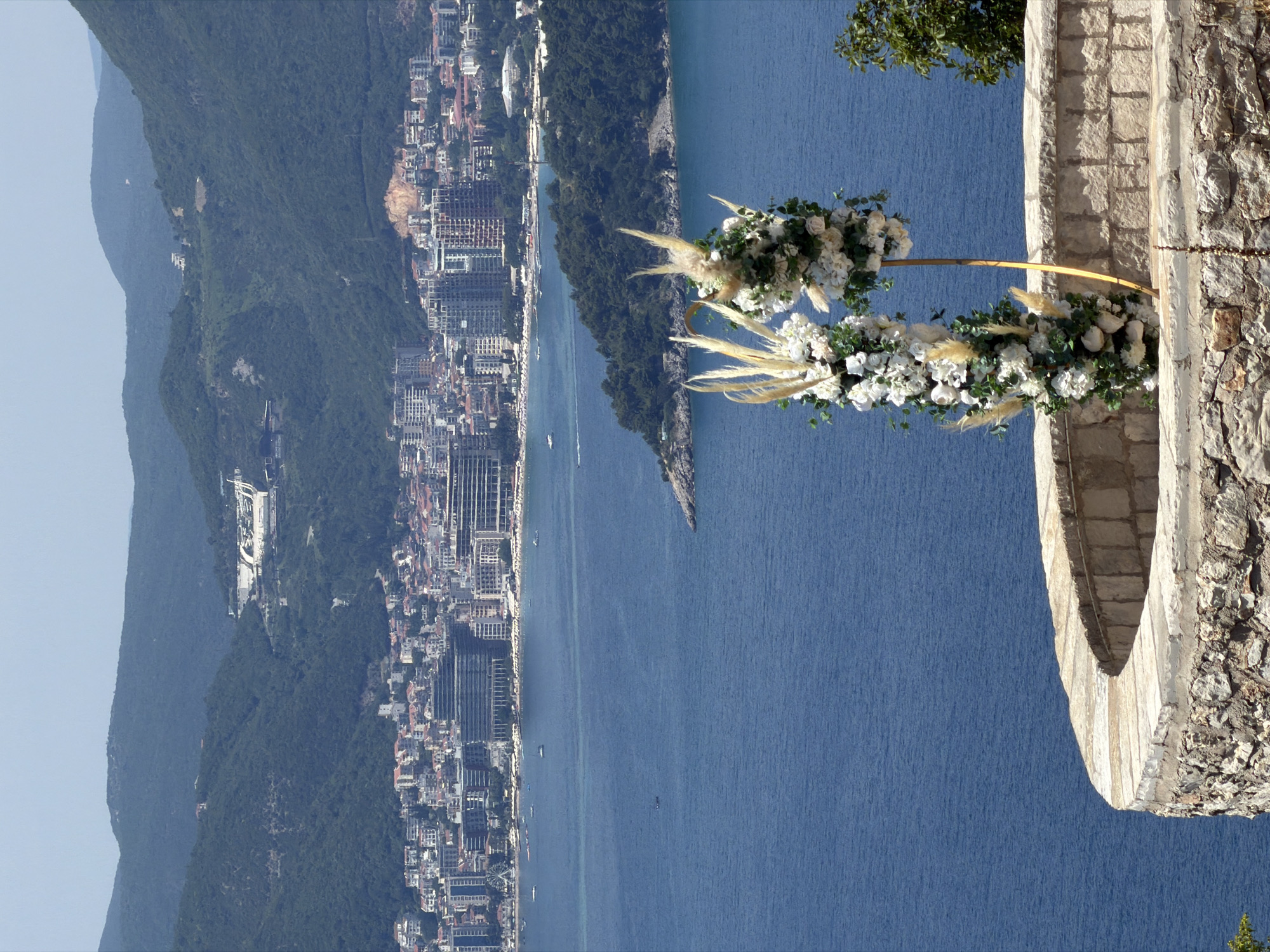 In the city of Sveti Stefan, Montenegro, a scenic overlook presents a panoramic view of the Adriatic Sea and a coastal city. The foreground is dominated by a curved stone wall, constructed from rough-hewn blocks and capped with smooth, pale stones. Two elaborate floral arrangements, composed of white roses and pampas grass, stand on thin golden poles, positioned on the wall's surface.
Beyond the wall, the deep blue of the sea stretches out to meet a densely populated coastline. A sprawling city, characterized by a mix of modern and older buildings with red-tiled roofs, climbs the foothills of verdant, tree-covered mountains. Buildings range from low-rise structures to taller, contemporary high-rises. Boats of various sizes dot the water, and a thin strip of beach, lined with umbrellas, is visible along the shore. Further inland, the mountains rise steeply, their slopes covered in a dense, dark green forest. The sky is clear and pale blue, indicating a bright, sunny day. The overall scene captures a serene coastal landscape with a touch of celebratory decor.