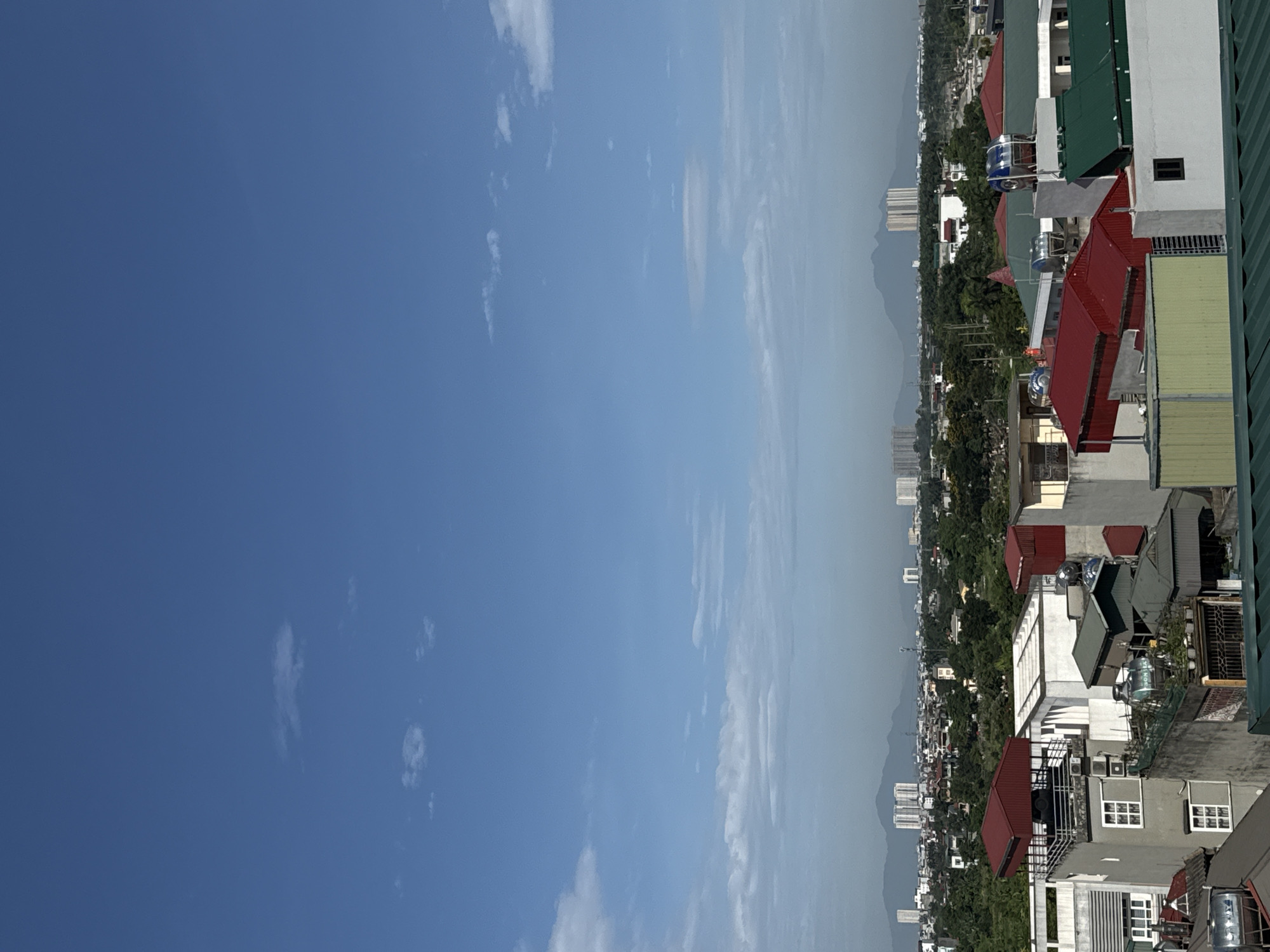 The image depicts a cityscape in Hà Nội, Vietnam, under a bright blue sky with scattered clouds. The foreground consists of rooftops of closely packed buildings, primarily composed of residential structures. The roofs are a mix of colors, including red, green, gray, and white. The buildings appear to be a mix of old and newer construction. In the background, there is a distant mountain range shrouded in a light haze. The photograph is vertically oriented.