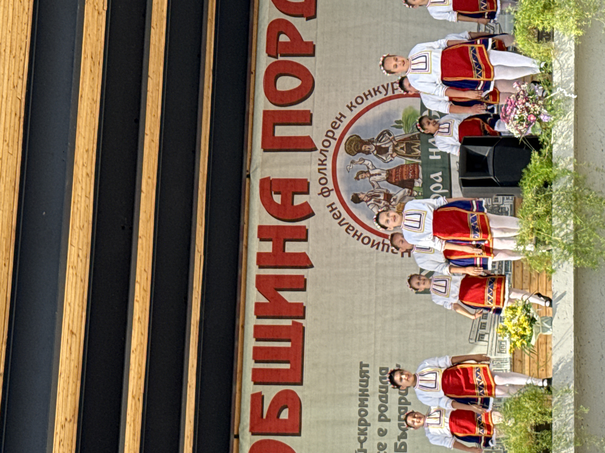 In Pordim, Bulgaria, a group of young girls in traditional Bulgarian folk costumes perform on a stage. The girls, with their hair adorned with floral wreaths, wear white blouses with intricate embroidery and red skirts with blue and gold trim. They are positioned across the stage, with some girls in the foreground and others behind them. A large banner with red Cyrillic lettering hangs behind them, partially obscured by the performers. The banner appears to say "ОБЩИНА ПОРОДИМ" (Municipality of Pordim) and "НАЦИОНАЛЕН ФОЛКЛОРЕН КОНКУРС" (National Folklore Competition). To the left of the banner, there is text in smaller font, and a circular emblem with depictions of traditional figures in folk attire. The stage itself is made of wood, with plants and a flower arrangement visible in the foreground. A black speaker is placed on the stage to the right of center. The backdrop suggests an outdoor event, with a wooden ceiling structure visible at the top of the image. The overall scene captures a cultural performance during a festival or competition in Pordim.
