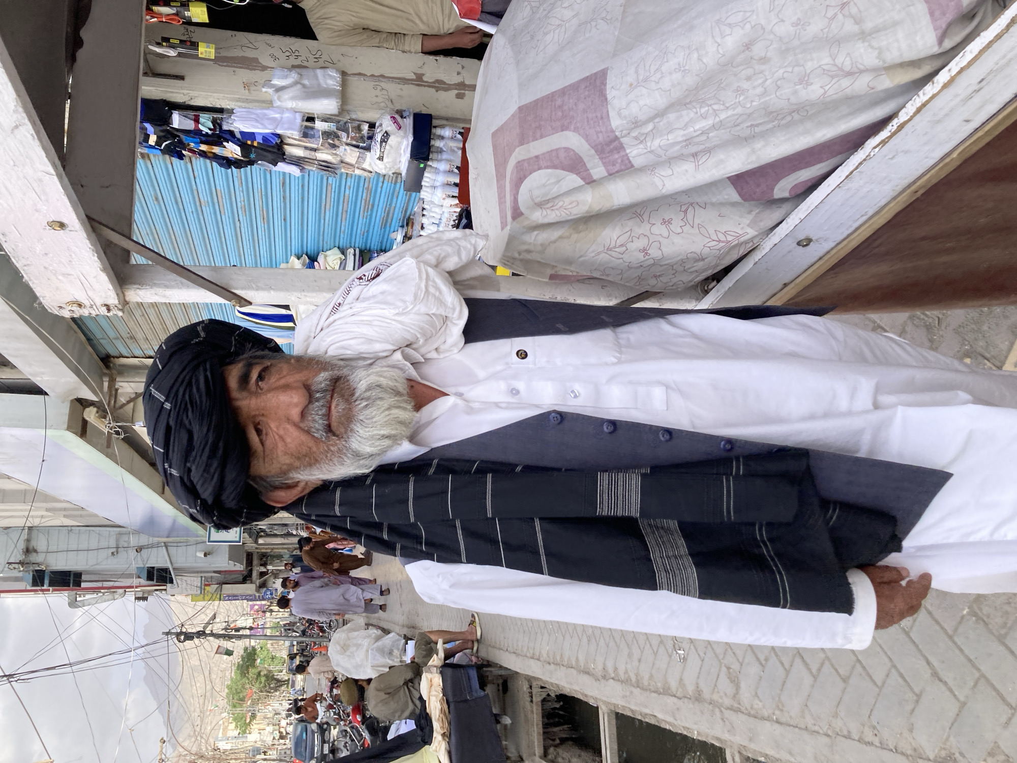 The image shows an elderly man with a grey beard, wearing a black turban and traditional Pakistani clothing, likely standing on a street in Quetta City Tehsil, Pakistan. He is positioned near a storefront with a blue roll-up door. The street scene includes other people, motorcycles, and vehicles. Power lines crisscross above the street. In the background, mountains are visible. A white cloth with a patterned design appears to be draped or displayed nearby.