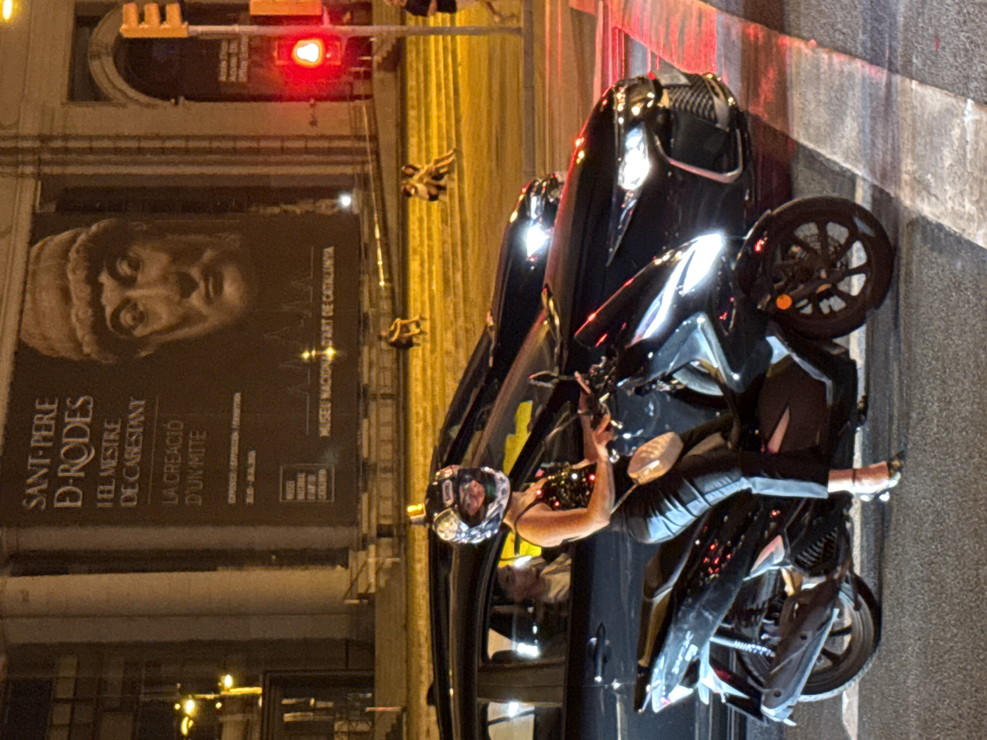 The image captures a street scene at night in Barcelona, Spain, likely near the Palau Nacional which houses the Museu Nacional d'Art de Catalunya. In the foreground, a woman wearing a black helmet with reflective accents and a sparkling black top is seated on a black scooter. She has on black pants and is wearing high heels. A light-colored purse is slung over her shoulder.
Behind her, a dark-colored car is stopped at a traffic light, with its headlights illuminated. The driver of this car, a person with dark hair, can be seen through the window. Another dark-colored car is visible further ahead, also stopped.
In the background, a large banner with text in Catalan is displayed on the side of a building. The text includes "SANT-PERE D'RODES," "EL MESTRE DE CABESTANY," and "LA CREACIÓ D'UN MITE." Below this, smaller text indicates "EXPOSICIO/EXPOSICION" and dates. The "MUSEU NACIONAL D'ART DE CATALUNYA" is also legible. Traffic lights, one of which is red, are visible, indicating the vehicles are stopped. Several people are seated on illuminated steps in the middle ground. The overall lighting suggests it is nighttime, with artificial lights illuminating the scene.