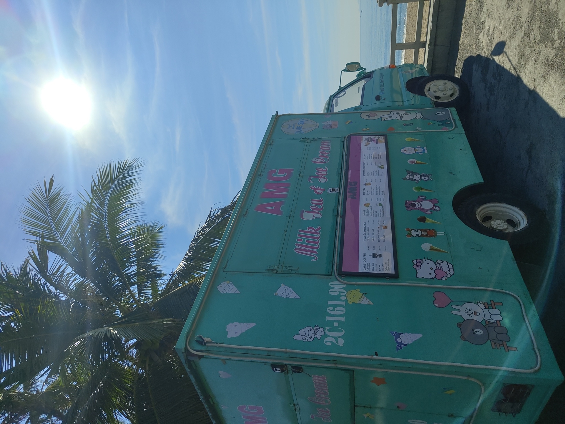 This is a medium shot, eye-level, outdoor, daytime, vertical photograph of a teal food truck that says "AMG Milk Tea & Ice Cream" on the side. The truck is parked on a paved surface with a railing and the ocean in the background. A palm tree is visible in the upper left corner, and the sun is shining brightly in the upper right corner. The truck is decorated with various cartoon characters and ice cream cones. There is a menu displayed on the side of the truck. The license plate on the truck reads "2C-161.90". The scene appears to be in Nha Trang Ward, Vietnam, due to the presence of palm trees, ocean, and what appear to be Vietnamese menu items. There are no people visible in the image.