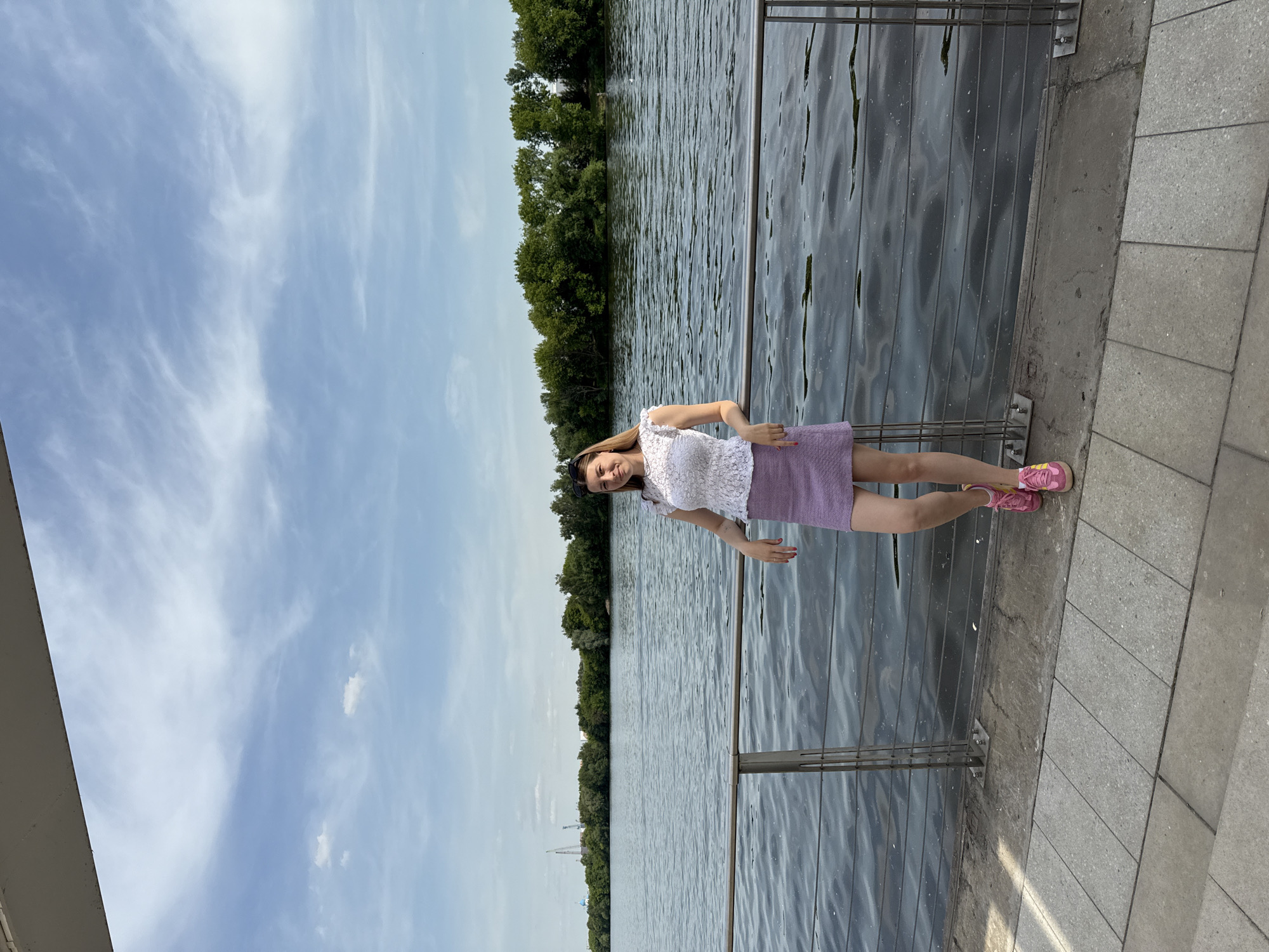 The image displays a woman standing on a paved embankment alongside a large body of water, identified as the Moskva River in Moscow, Russia. The scene is set under natural daylight, suggesting late morning or early afternoon, beneath a blue sky with scattered white, wispy clouds and some smaller cumulus formations.
The central subject is a woman, appearing to be in her 20s or 30s, with long, straight brown hair. She is wearing a white, short-sleeved top with a textured, possibly lace, pattern, and a light purple mini-skirt. Pink and yellow athletic sneakers complete her attire. Sunglasses are perched atop her head. She stands with her left arm casually resting on a metal railing that spans the length of the embankment. Her right hand is positioned near her left elbow. The woman exhibits a subtle smile and is looking directly towards the camera.
The immediate foreground features grey, rectangular paving slabs forming the walkway. A modern, two-tiered metal railing, comprising a thicker top rail and several thinner horizontal bars, runs parallel to the river's edge, separating the paved area from the water. The water itself is dark blue-grey, exhibiting numerous small ripples across its surface, indicative of a slight current or wind.
The background consists of a dense line of mature green trees on the opposite bank of the river. Above the tree line on the far left, several indistinct industrial structures, including a blue dome and multiple tall cranes, are visible against the sky. The horizon is clearly defined by the meeting point of the water, the trees, and the sky.