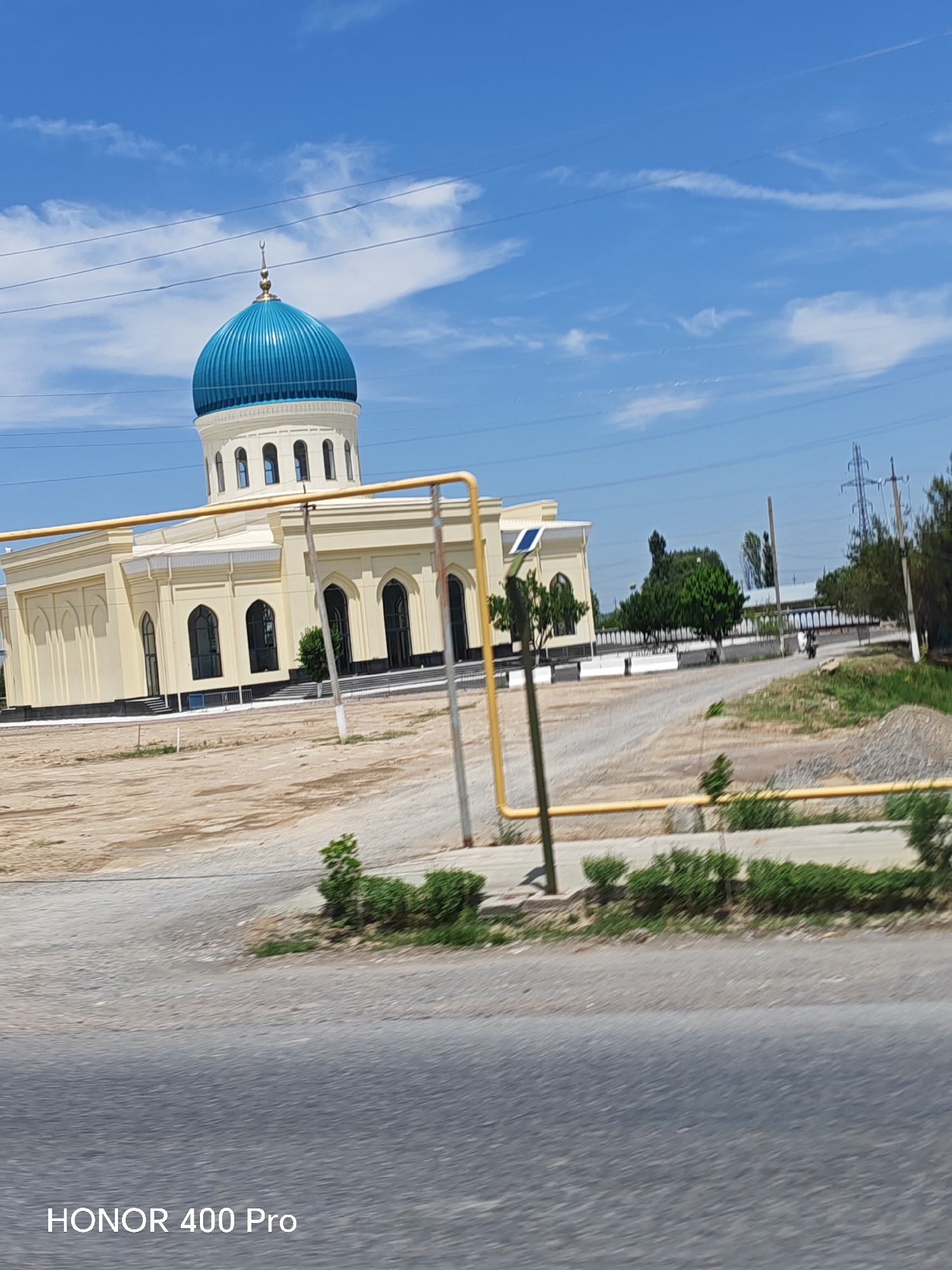 This image captures a mosque with a prominent blue dome in Sardoba, Uzbekistan. The mosque is a light yellow color with arched windows and a white trim. In front of the mosque, there is a yellow pipe structure. The area surrounding the mosque appears to be a dusty, unpaved lot with some scattered greenery and trees in the background. Power lines and a utility pole are visible in the sky. The overall scene suggests a sunny day.
