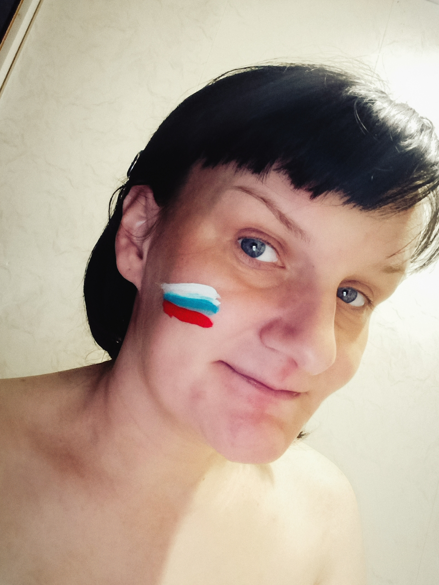 A close-up, head-and-shoulders portrait captures a woman indoors, potentially in Фоки, Russia. She has short, dark hair with bangs and bright blue eyes, looking directly at the camera with a subtle, gentle smile. On her left cheek, face paint in horizontal stripes of white, blue, and red is visible, strongly resembling the Russian flag. Her bare shoulders are visible, suggesting she is shirtless or wearing a strapless garment. The background is a plain, light-colored wall with a faint texture. The lighting is bright, illuminating her face. The image depicts a moment of personal expression, possibly showing national pride or support. No text is visible in the image.