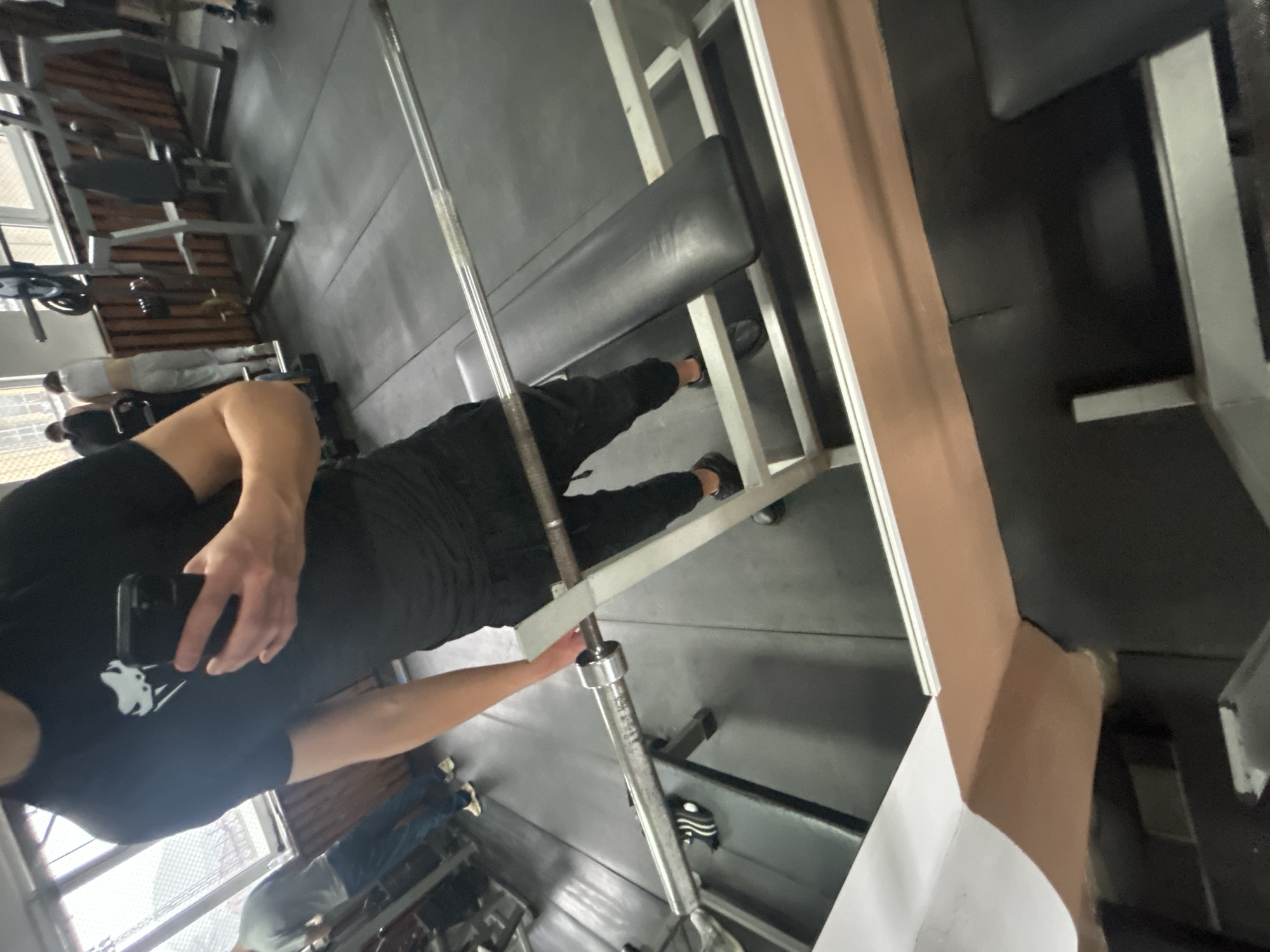 A person wearing black athletic clothing stands in a gym holding a smartphone and facing a weight bench with a barbell. The gym is located in Zlatograd, Bulgaria. Gym equipment is visible.
