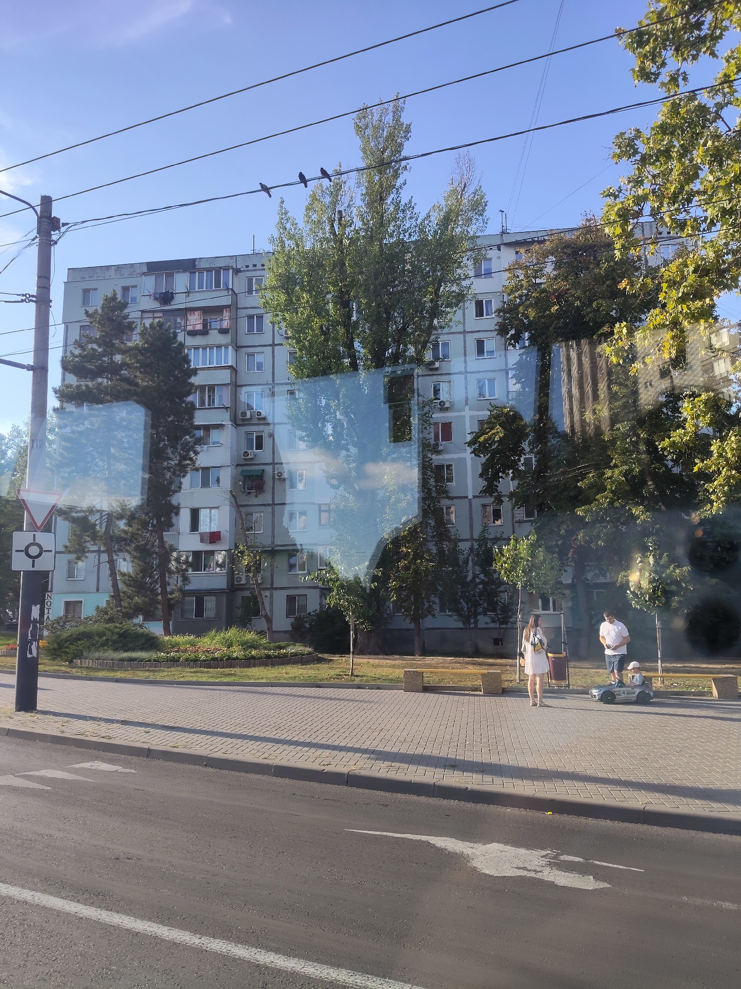 In Durlești, Moldova, this image captures a sunny day scene with an apartment building and a street. The building, a multi-story structure with a light blue facade, features numerous windows, some with air conditioning units and clothes drying on balconies. The building's exterior shows signs of wear. In front of the building, trees and green spaces are visible, including a circular flower bed. A traffic sign with a red triangle and a circle is mounted on a pole on the left side of the image. The foreground shows a paved sidewalk next to a road with lane markings. On the sidewalk, two adults and a child are present. A woman in a white dress stands near a trash can, while a man in a white polo shirt and blue shorts stands next to a small, toy electric car occupied by a child. Two wooden benches are placed on the sidewalk. The scene is framed by power lines overhead and a reflection of trees and the sky on a glass surface, possibly from a vehicle window.