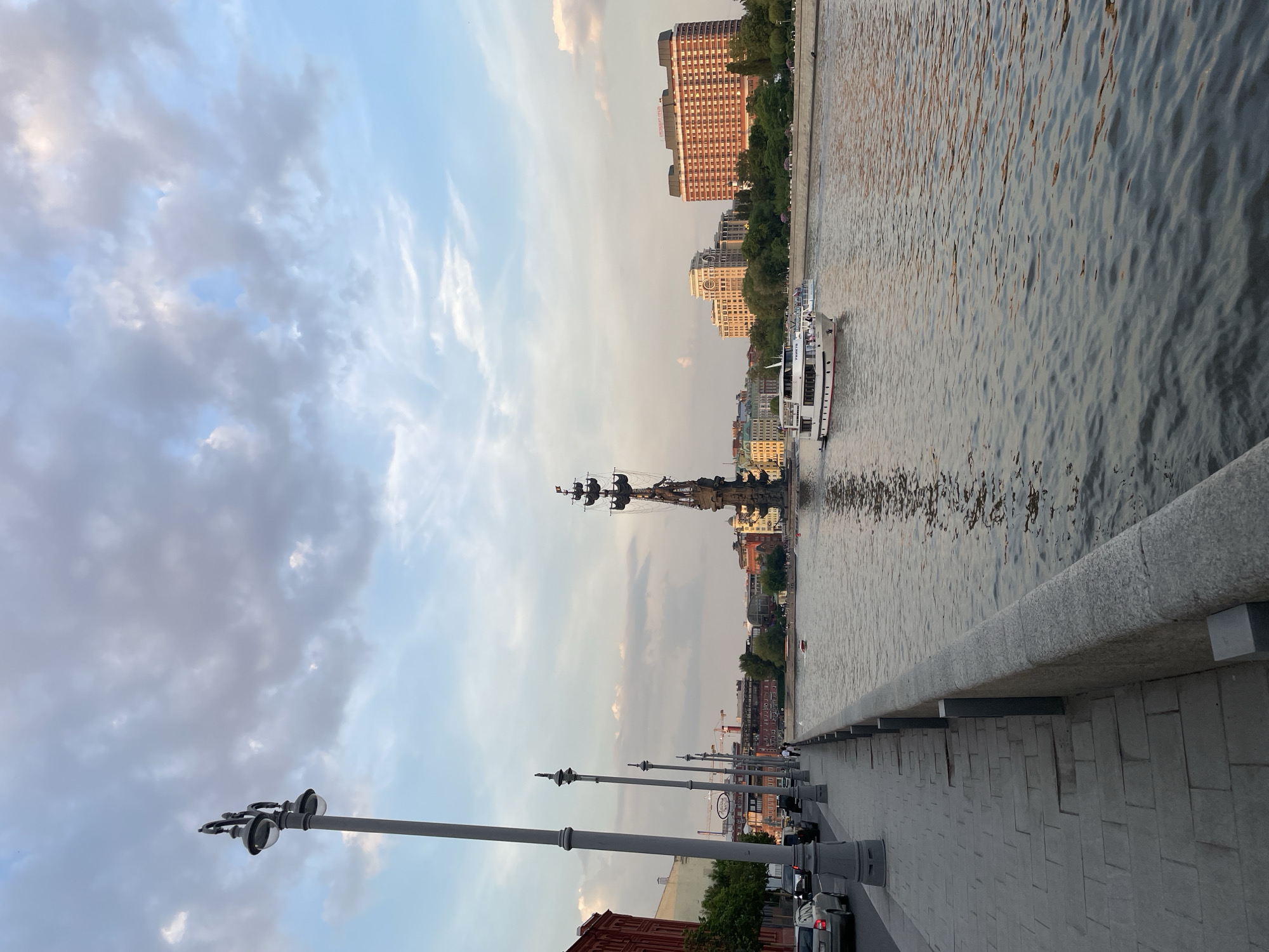 The image shows a view of the Moskva River in Moscow, Russia, under a cloudy sky. In the foreground, there is a stone embankment with streetlights lining the path. Cars are parked alongside the road. The river reflects the city skyline. A tour boat is seen on the river. The Peter the Great Statue is visible in the river. Buildings of varying heights and architectural styles line the opposite bank.