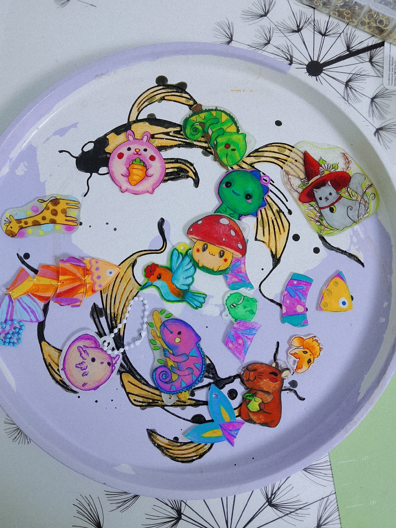 A top-down shot shows a light lavender plate with a hand-drawn black and gold koi fish design. Various colorful, cartoonish charms and trinkets are scattered on the plate, including: a pink bunny holding a carrot, a green chameleon, a green dinosaur, a mushroom with a face, a hummingbird, a giraffe, various fish with rainbow segments, a purple axolotl, a blue and purple chameleon, a squirrel holding an apple, a fox face, a cat wearing a red hat with flowers, and some geometric segments. A chain of small white beads is also present. In the top right corner, a container with metal jump rings is visible, and part of a dandelion illustration is on the edge of the plate. The background includes a white surface with black dandelion designs and a light green surface. The location is unknown and cannot be determined to be Chaikovsky, Russia based on this image alone.