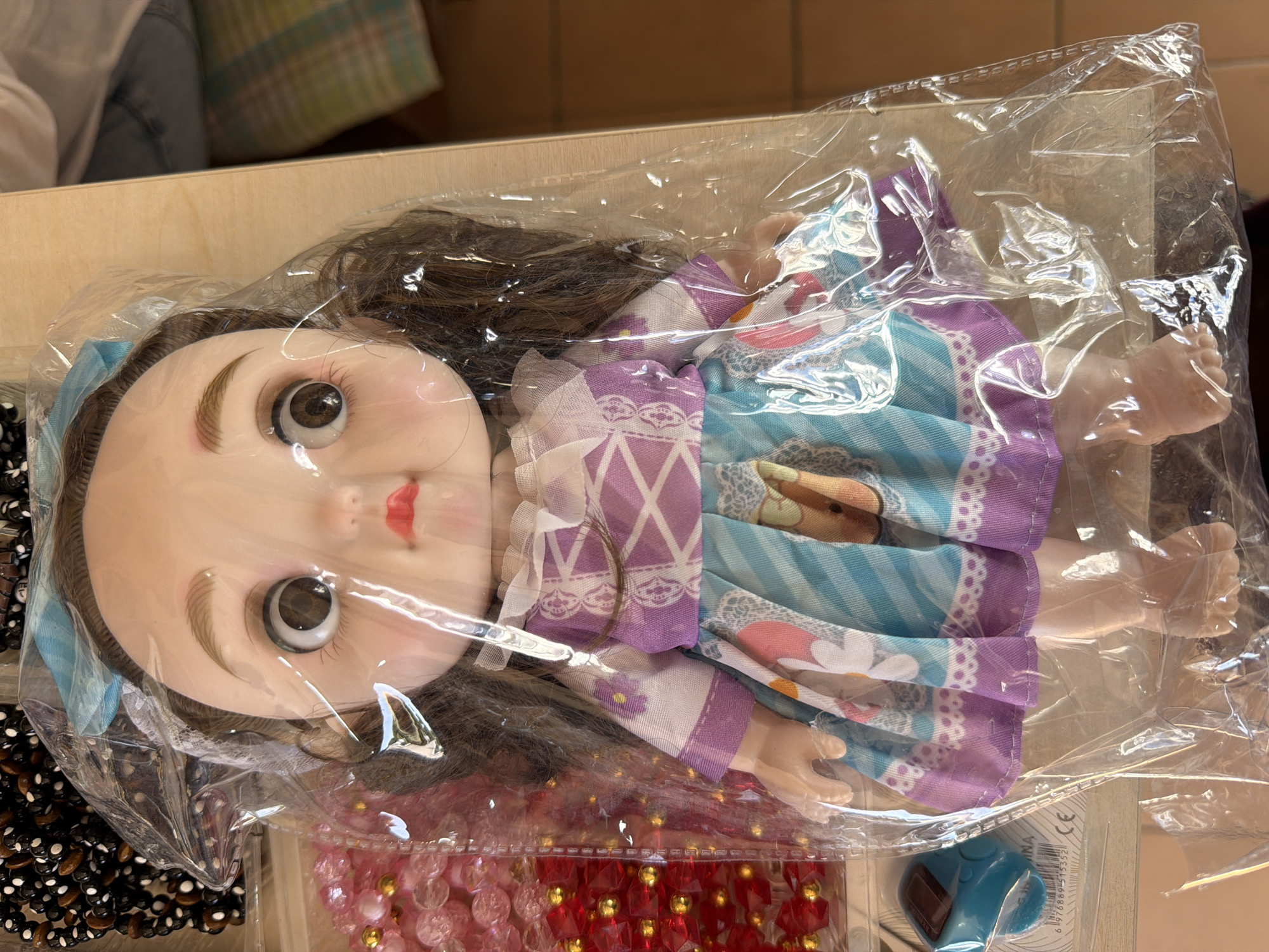 A doll is encased in clear plastic packaging and is positioned on a wooden surface. The doll has brown hair, large brown eyes, and is wearing a purple and white dress with a patterned skirt. To the left of the doll, there are various strands of beads, some pink and some red with gold accents. A small blue object with a digital display is also visible to the left. In the background, on the right side of the image, there is a blurred view of a checkered fabric. The scene is illuminated by natural light, casting soft shadows. The location of Zangiata, Uzbekistan is not discernible from the visual information provided in the image.