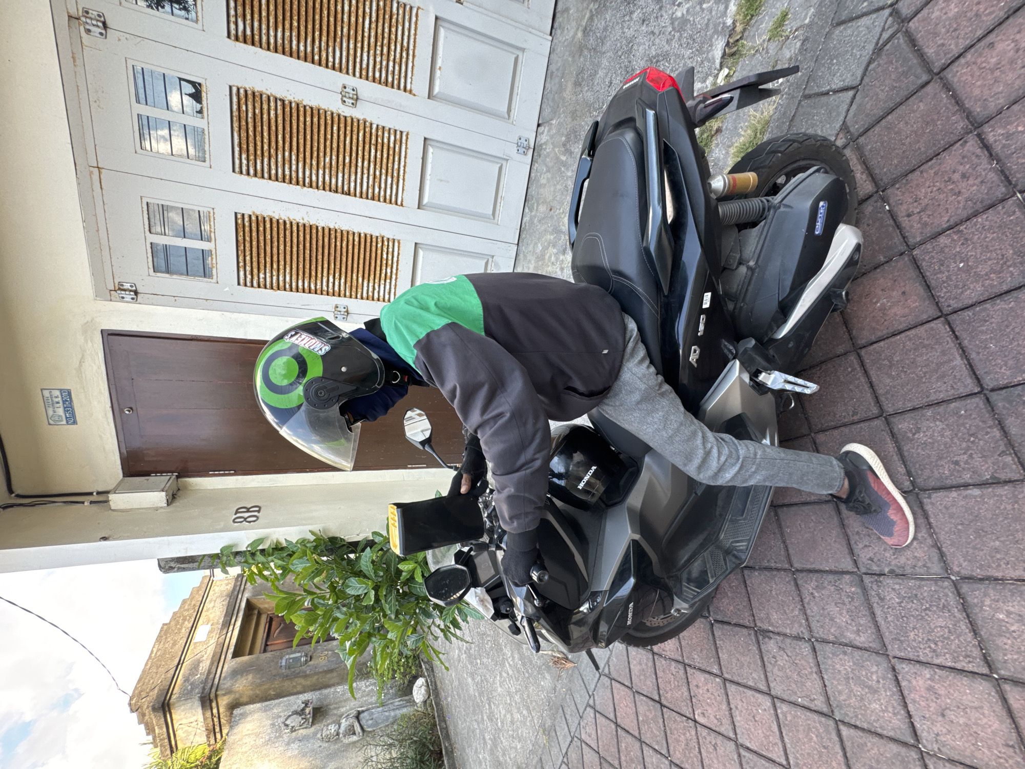 A person is depicted on a black Honda scooter, seemingly preparing for a ride or having just arrived. They are wearing a black and green jacket, grey pants, black gloves, and a black and green helmet with "SHOPEEFOOD" branding visible on the side. Their hands are placed on the scooter's handlebars.
The scene is an outdoor residential area during the daytime, under what appears to be sunny weather, judging by the bright reflections in the windows. The scooter is parked on a reddish-brown paver sidewalk. Adjacent to the scooter is a building with a large, light-colored metal garage door that shows significant rust, featuring small rectangular windows and horizontal slatted panels. Next to the garage door is a smaller, dark brown wooden door. Above this door, a small plaque reads "NO. M53/H.2016", and the number "8B" is visible on the wall below it. Green foliage is visible in the foreground on the left side. The scooter itself has "HONDA" logos visible on the front and side panels. The location context provided is Kuta, Indonesia.