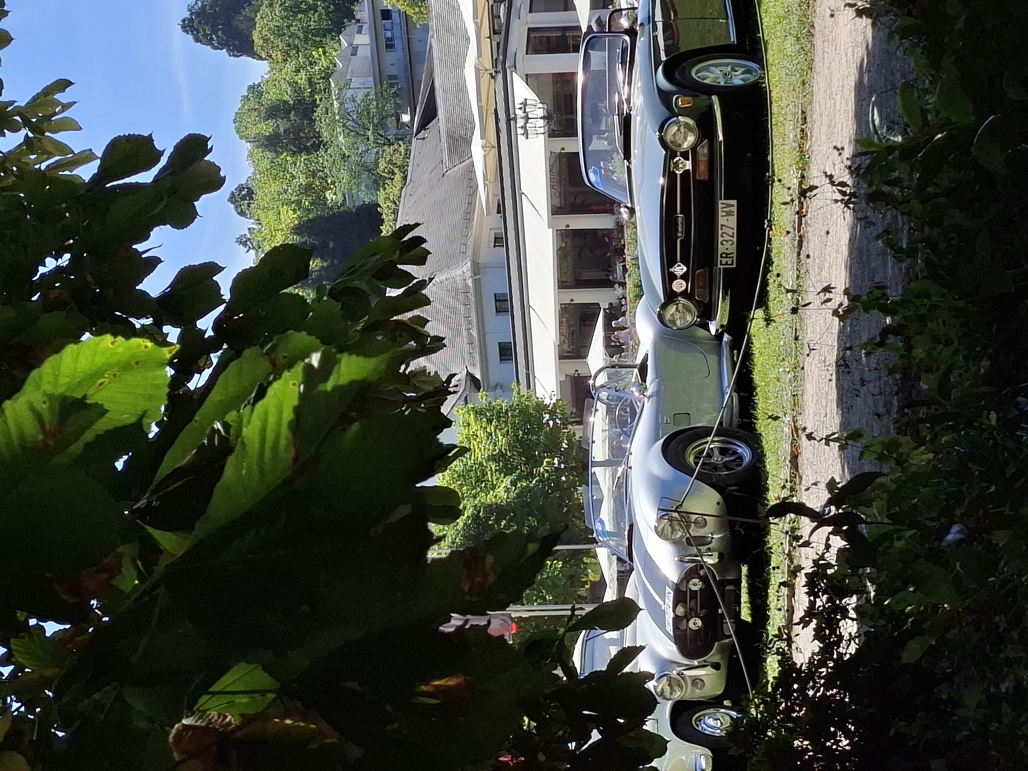 The image captures a sunny outdoor scene in Baden-Baden, Germany, likely during a car show or event. In the foreground, large green leaves of a tree partially obscure the view. Visible in the middle ground are two classic sports cars. The car on the left is a silver convertible with black racing stripes, featuring large round headlights and a chrome bumper. To its right is a dark green convertible, also with round headlights and a chrome bumper. This car has a license plate that reads "ER-327-WV." Behind the cars, a building with a large white awning and outdoor seating areas is visible. Further in the background, a lush green hillside with trees and more buildings can be seen under a clear blue sky. The ground in front of the cars appears to be a mix of grass and gravel.