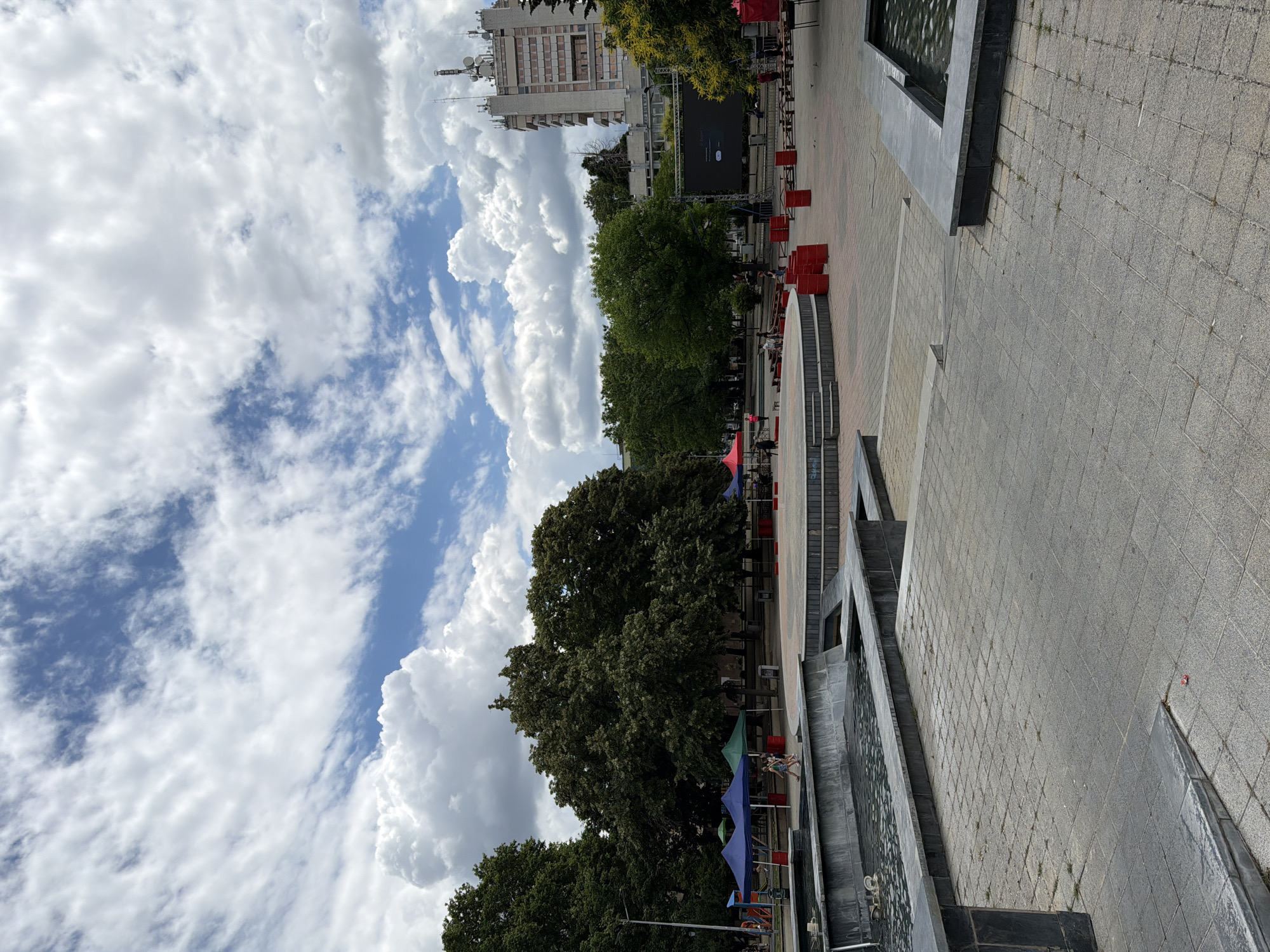 The image shows a public square in Pazardzhik, Bulgaria, on a cloudy day. In the foreground, a modern concrete fountain with water is visible, with patterned paving stones surrounding it. Further back, there is a raised circular stage area with red cylindrical objects scattered around it. Several tents with blue and green canopies are set up to the left, possibly for a market or event. A large screen is set up on the right side of the square, suggesting an outdoor performance or viewing area. Behind the square, a tall, light-colored building with many windows and communication antennas on its roof dominates the skyline. Lush green trees are interspersed throughout the scene, providing shade. A few people are present in the square, some walking and others appearing to be setting up or attending to the tents. The overall impression is a vibrant public space, possibly preparing for or hosting an event.