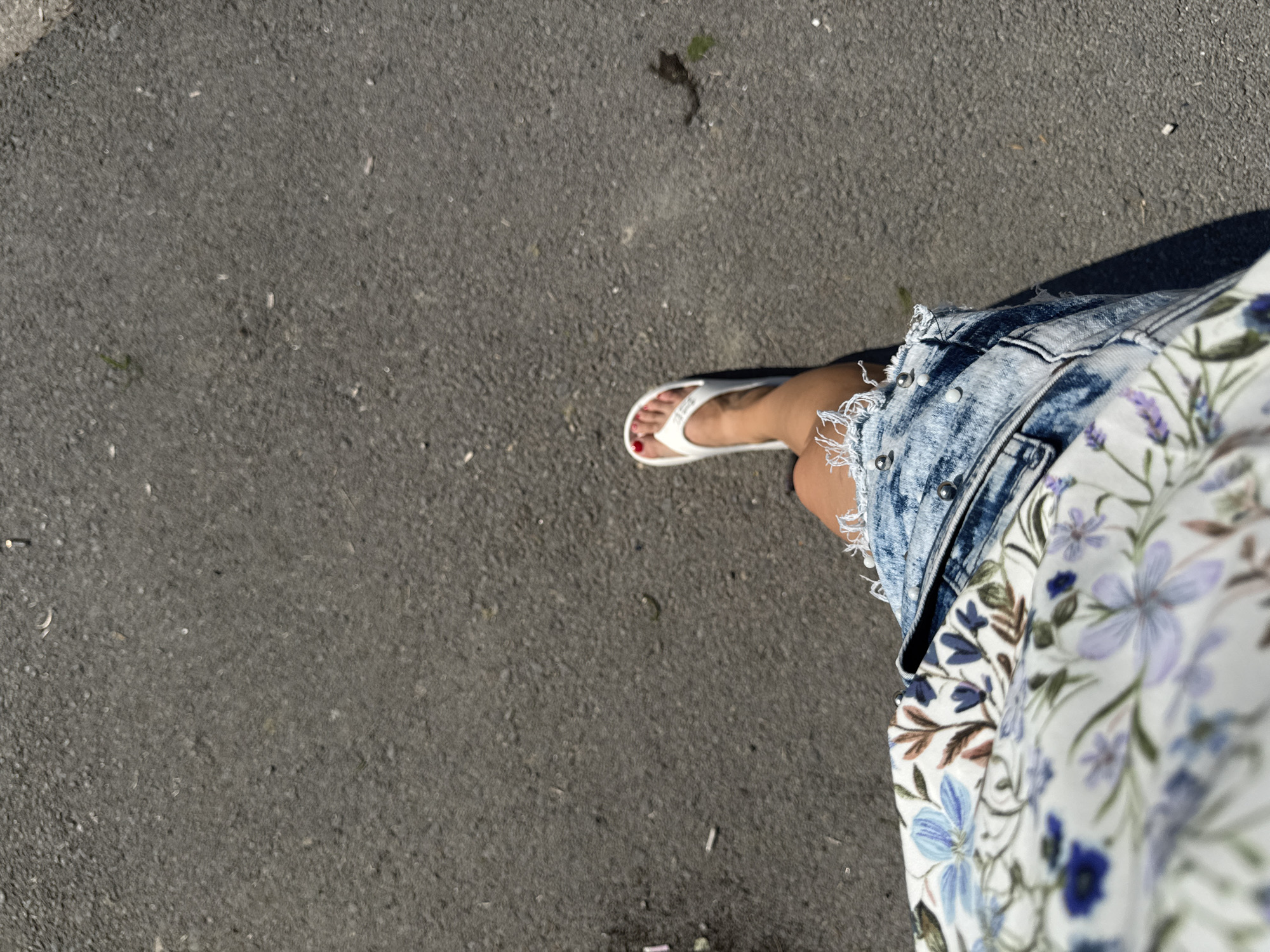 A first-person perspective shot captures a person's legs and feet walking on an asphalt surface. The individual is wearing white flip-flops with red-painted toenails and a distressed denim skirt adorned with silver studs. A floral-patterned fabric, possibly part of a shirt or dress, is visible in the lower left portion of the frame, featuring blue, purple, and brown floral motifs against a white background. The asphalt is dark gray with scattered small debris. The lighting suggests it is daytime with direct sunlight casting a shadow to the left of the visible leg. The location is unspecified, but the environment appears to be an outdoor urban or suburban setting.