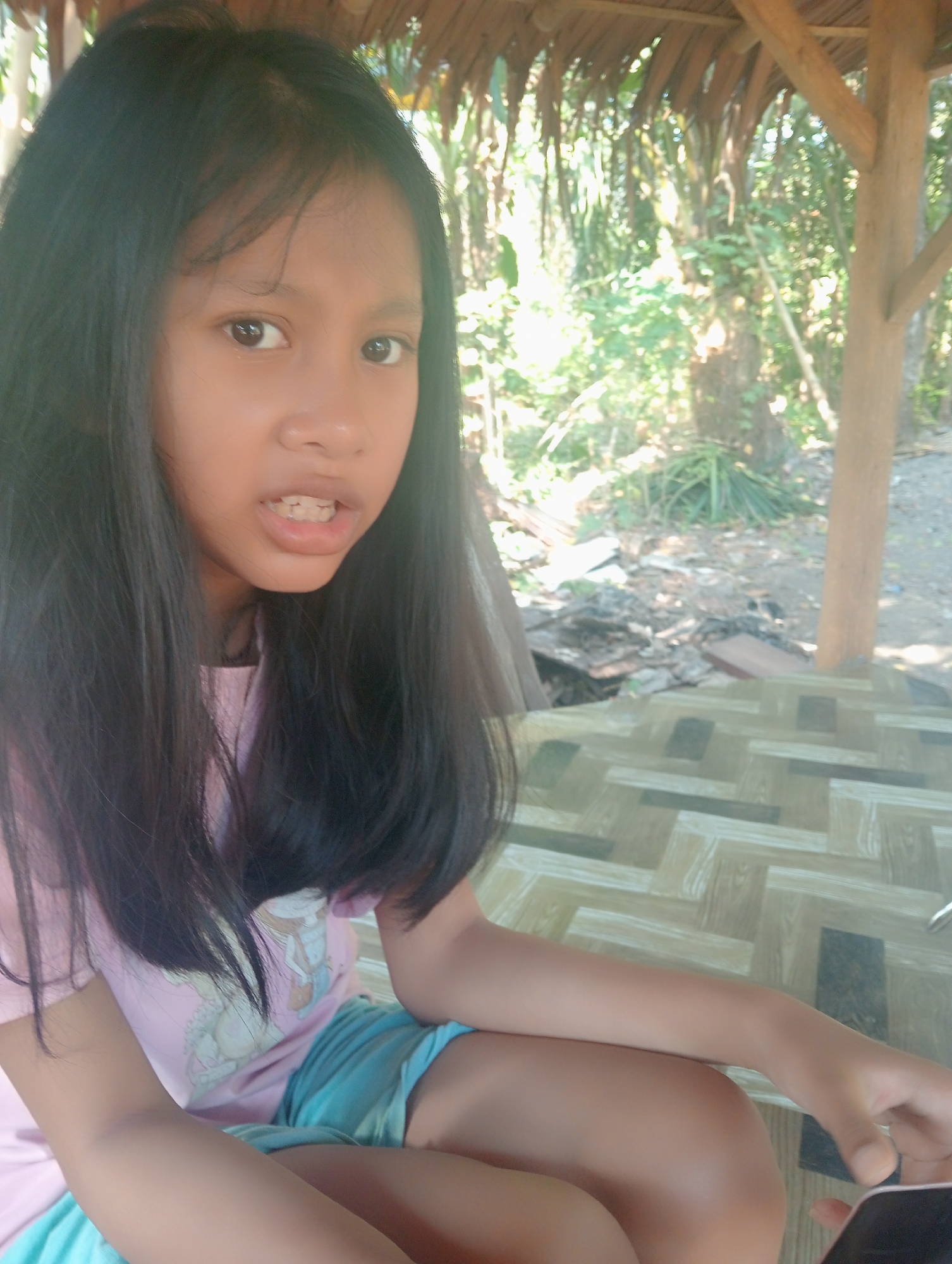 A young girl with long, dark hair and fair skin is pictured sitting outdoors under a thatched roof structure. She is wearing a pink t-shirt with a graphic print and blue shorts. Her mouth is slightly open, revealing her teeth. The background shows lush greenery and trees, suggesting a rural or natural setting. The surface she is sitting on appears to be a patterned mat or flooring. The location is identified as Muara, Indonesia.
