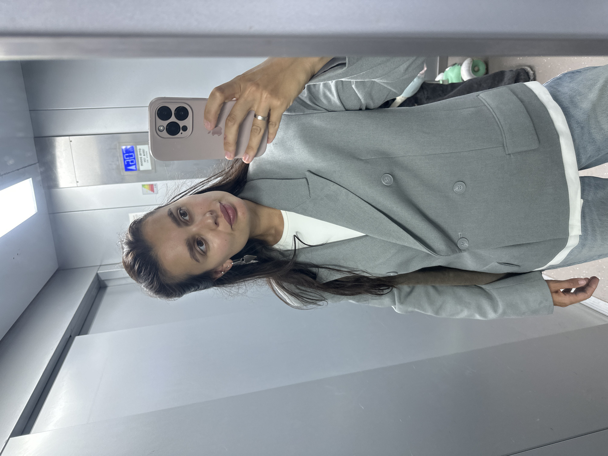 A woman stands in an elevator, taking a mirror selfie. She is wearing a grey double-breasted blazer over a white t-shirt and blue jeans. She has long, dark hair pulled back from her face and is wearing a silver ring on her right hand and dangling earrings. The elevator walls are grey, and a digital display on the elevator panel shows the number "3" in blue. The location is an elevator in an apartment building in Chaikovsky, Russia.