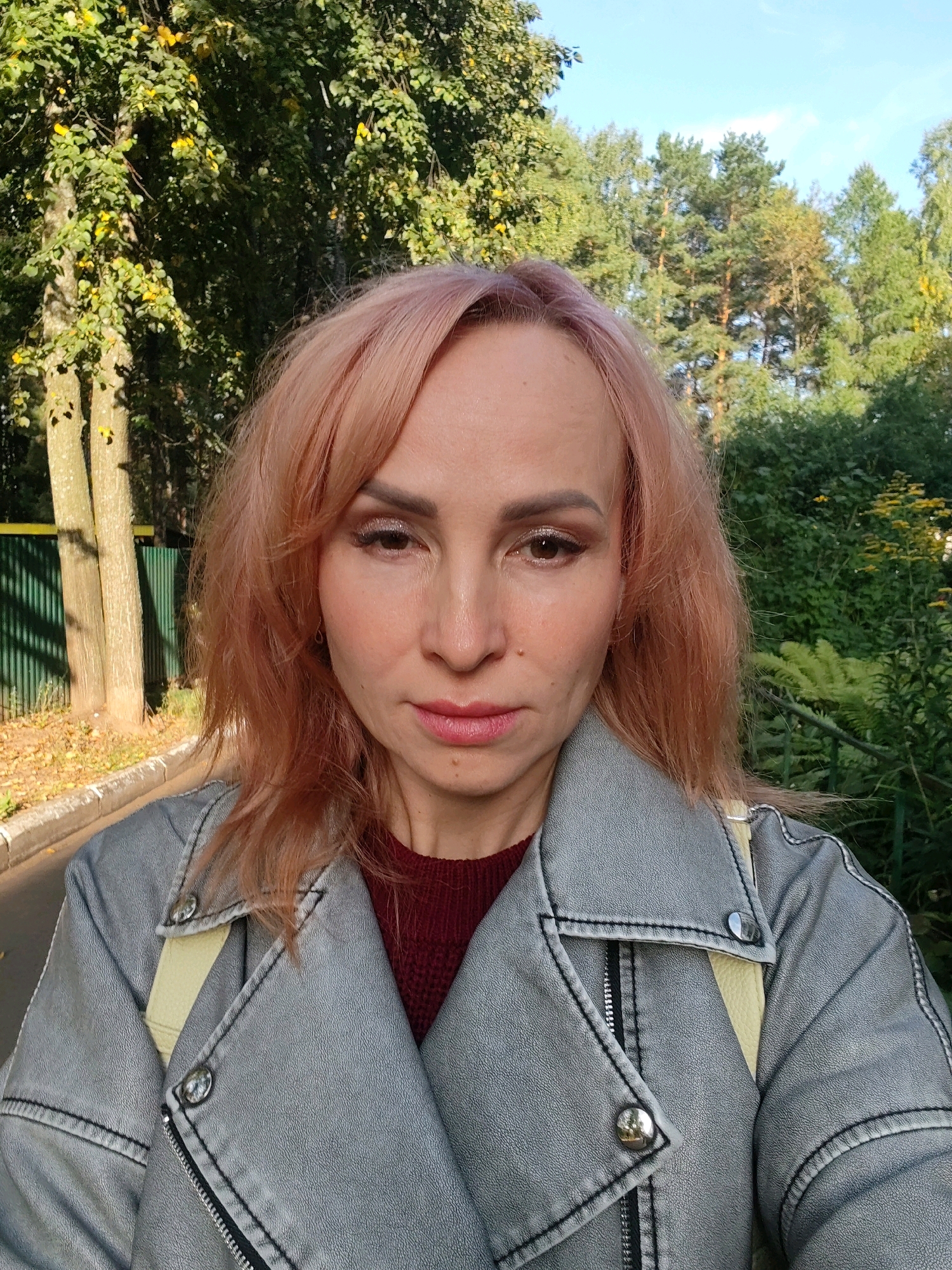 A medium shot captures a woman with light pink hair and light-colored makeup, wearing a gray jacket over a maroon sweater. She is outdoors, with trees and foliage in the background, suggesting a park or natural area. The lighting indicates it's daytime, possibly late afternoon due to the warm tones. The woman's expression is neutral, with her gaze directed towards the camera. The scene appears to be a casual, outdoor setting in Chaikovsky, Russia.