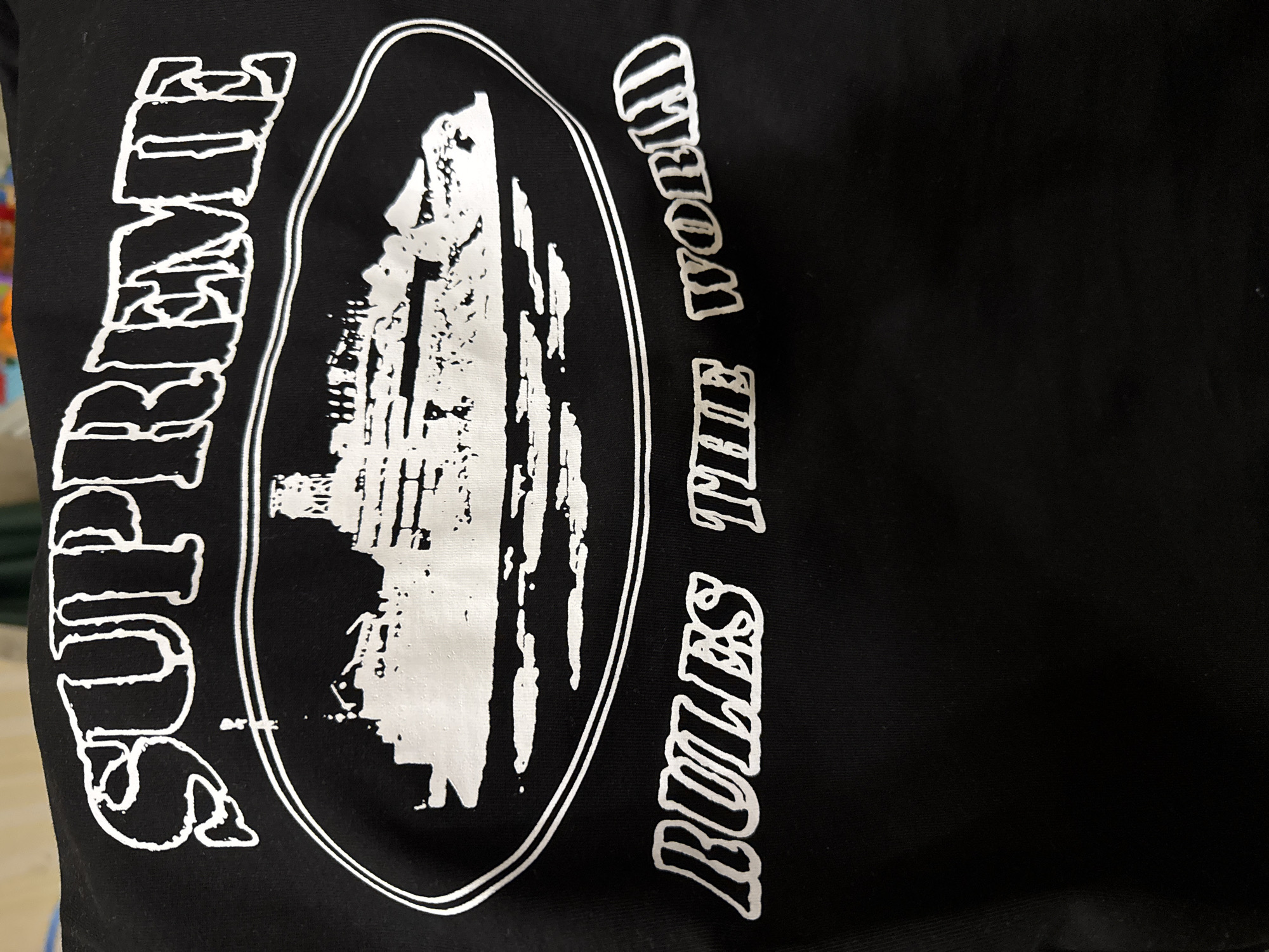 The image shows a black fabric, possibly a garment, with a white graphic design. The design features the word "SUPREME" vertically on the left, an oval shape in the center containing a white graphic of what appears to be a building or structure. To the right of the oval, the phrase "RULES THE WORLD" is printed vertically. The overall style is bold and graphic. There is no indication in the image that it is taken in Yerevan, Armenia or any other particular location.