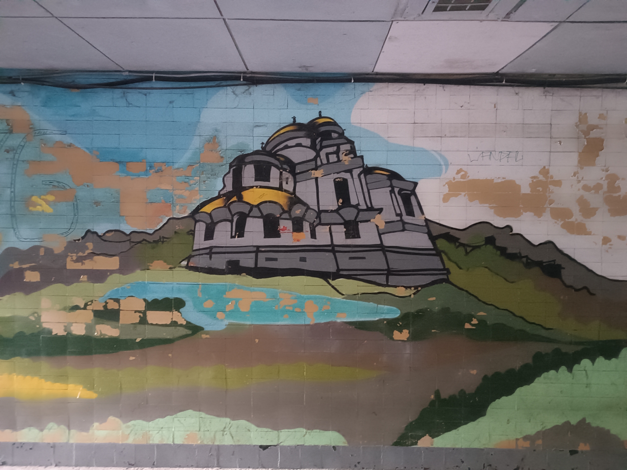 This is a photograph of a mural painted on a tiled wall, likely in an urban underpass or tunnel. The mural depicts a stylized landscape with a large, grey and yellow domed structure resembling a cathedral or church dominating the center. Rolling hills in shades of green and brown surround the structure, with a blue body of water in the foreground. The sky is painted in blue with abstract cloud shapes in lighter blues and orange-brown patches, suggesting peeling paint or decay of the underlying wall. The wall itself is made of rectangular tiles, some of which are chipped or missing, revealing the underlying material. The ceiling above the mural is a white, tiled grid with electrical conduits running along it. Some faint graffiti appears to be present on the upper right portion of the wall. The overall impression is of an art installation in a somewhat neglected public space. The presence of a specific building or landmark within Novocherkassk cannot be determined from the image alone.