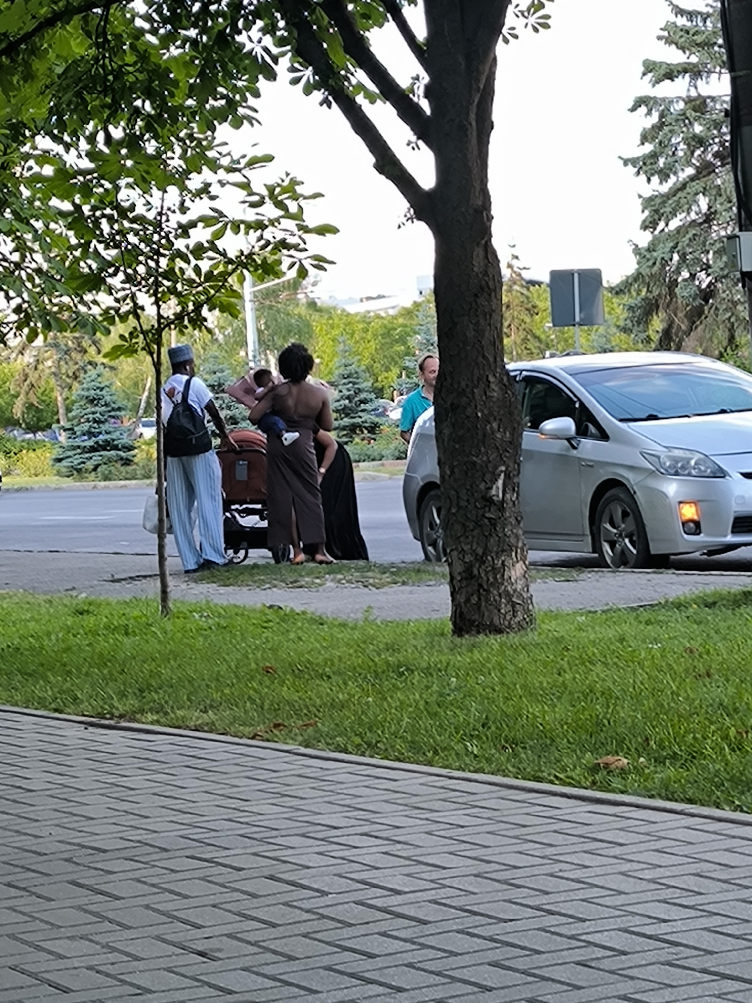 In Chișinău, Moldova, on a sidewalk next to a road, a group of people stand near a brown cart. A person wearing a white t-shirt and light blue pants, with a backpack, is near the cart. A woman with dark, curly hair is holding a baby and wearing a brown garment. Another person, mostly obscured by the woman, is wearing a dark garment. A silver car is parked on the road beside them. A man wearing a blue shirt is standing to the left of the car, partially visible. The environment includes trees and green grass.