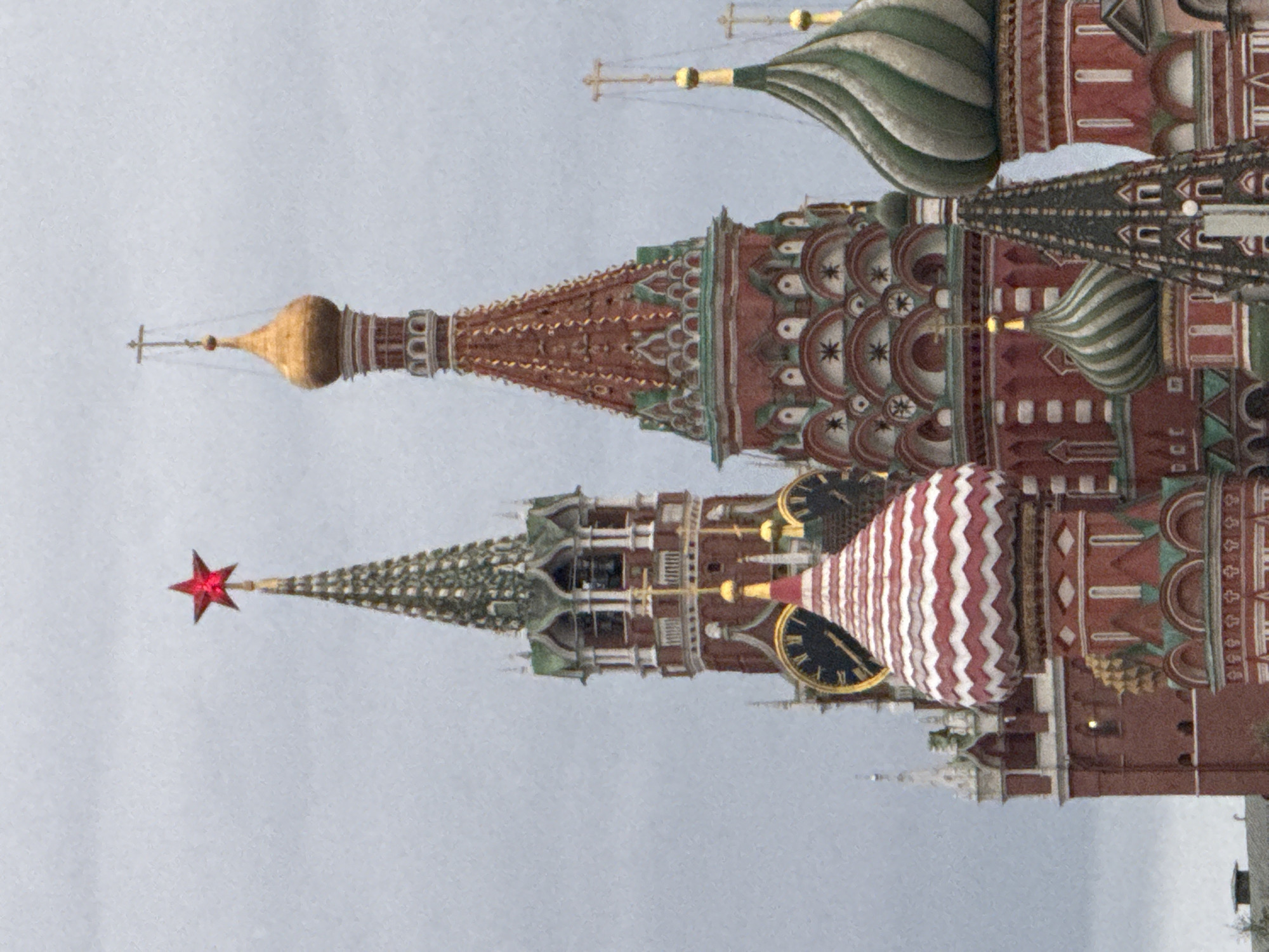 The image displays a close-up view of several onion domes and spires of Saint Basil's Cathedral in Moscow, Russia, under a cloudy sky. The most prominent structures are the red and white striped dome in the foreground, a large red tower with a green-roofed spire and a red star on top, and a tall, slender tower with a golden dome and a cross. To the right, other domes with green and white stripes and golden crosses are visible. The architecture features intricate patterns, arches, and colorful facades. The overall lighting is diffuse due to the overcast sky.
