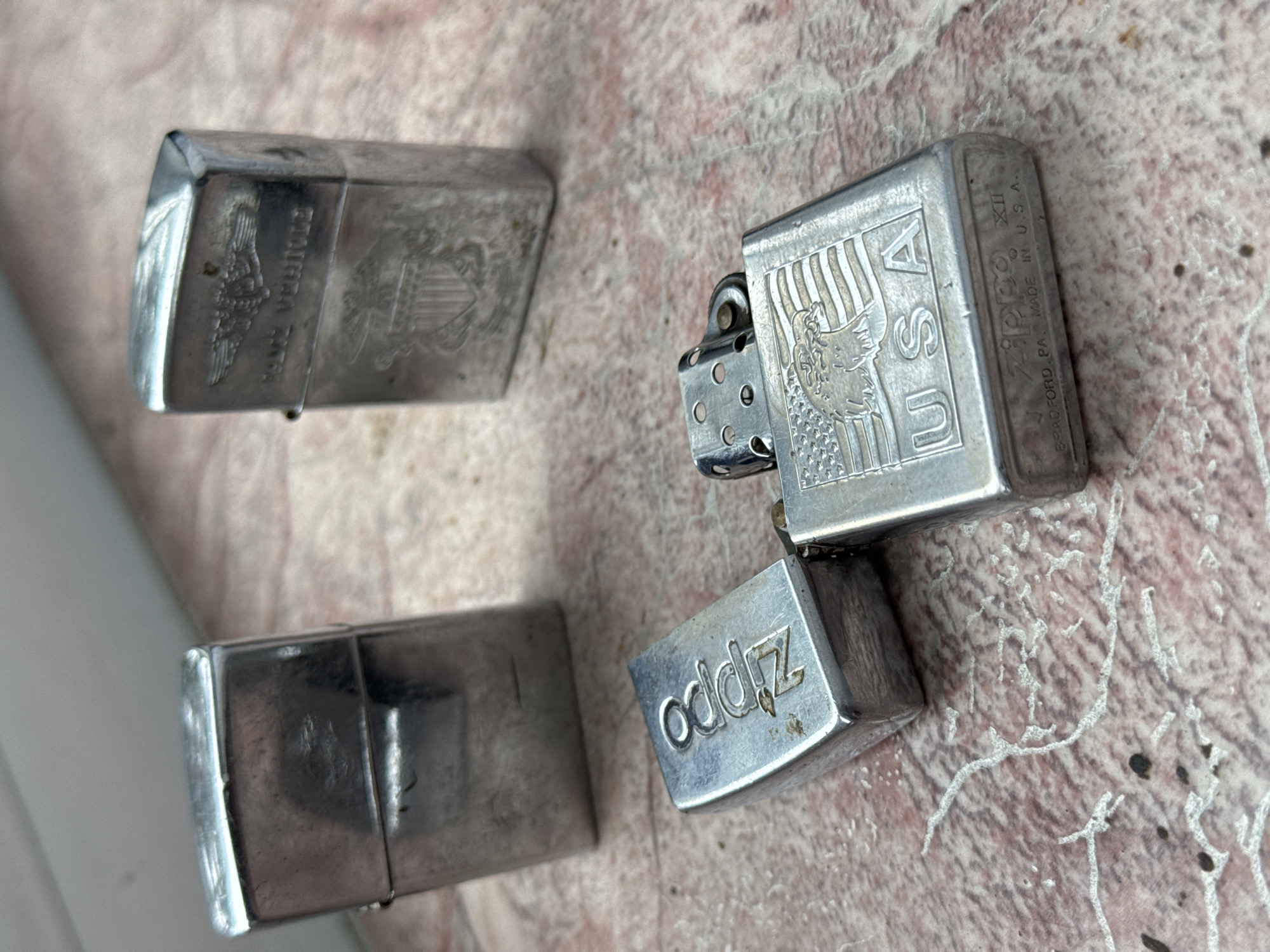 The image shows four Zippo lighters on a surface with a mottled pattern. One Zippo is open, revealing the inner workings and the name "Zippo" on the side. Another has an American eagle design with "USA." A third lighter has an eagle design and a partial inscription that appears to read, "POLICE MEMORIAL." The fourth Zippo is undecorated. Chuprino, Russia is not involved.
