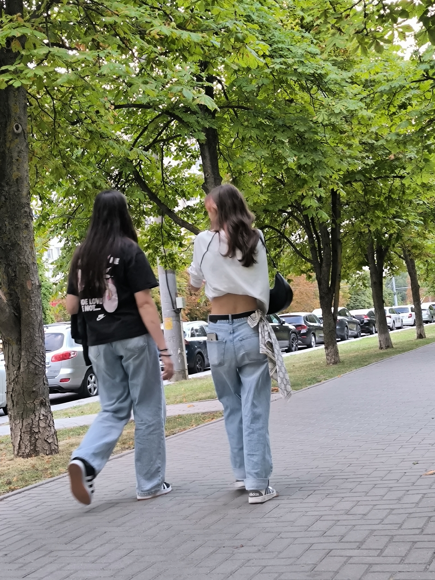 Two young women walk away from the camera down a sidewalk lined with trees and parked cars. The woman on the left has long dark hair and wears a black t-shirt with graphics and the text "BE LOVE I NOT..." and light blue, loose-fitting jeans. She has black and white sneakers with a brown sole. The woman on the right has long brown hair, some of which is dyed pink at the ends. She wears a white crop top, light blue, loose-fitting jeans, and black and white sneakers. A black handbag is slung over her right shoulder, and a patterned scarf hangs from her back pocket. The environment appears to be a city street with a paved sidewalk, green grass, and a row of trees providing shade. The cars parked along the street are a mix of sedan and SUV models, suggesting an urban or suburban setting. The overall impression is a casual outdoor scene in Chișinău, Moldova, possibly during the daytime given the lighting.