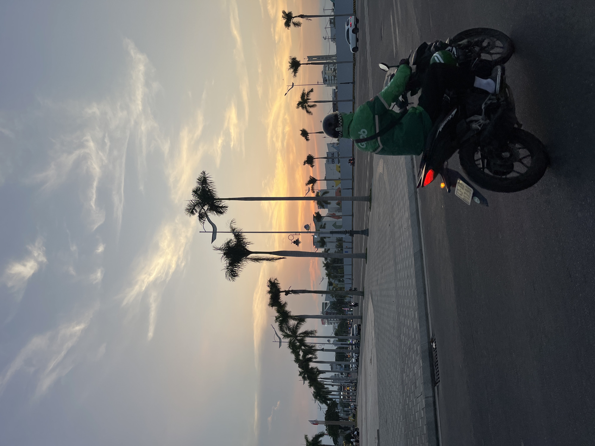 This portrait-oriented image captures an urban street scene in Da Nang, Vietnam, during either sunrise or sunset, indicated by the warm, golden light across the sky. The horizon glows with soft orange and yellow tones, gradually transitioning to a lighter blue with delicate, wispy white clouds higher up.
In the mid-ground, a person is seen on a dark motorcycle, positioned on the paved road adjacent to a wide, tiled sidewalk. The individual is wearing a bright green uniform jacket, prominently displaying the "Grab" logo on the back, and a dark helmet. The motorcycle's red taillight is illuminated.
A long line of tall palm trees frames the street, running parallel to the sidewalk and extending into the distance. Beyond these trees and across the street, various multi-story buildings and structures of an urban environment are visible, some with faint lights or signs. The scene depicts a typical moment of daily life in a tropical city during the 'golden hour', blending natural elements like palm trees and a vibrant sky with modern urban infrastructure and a local delivery service.