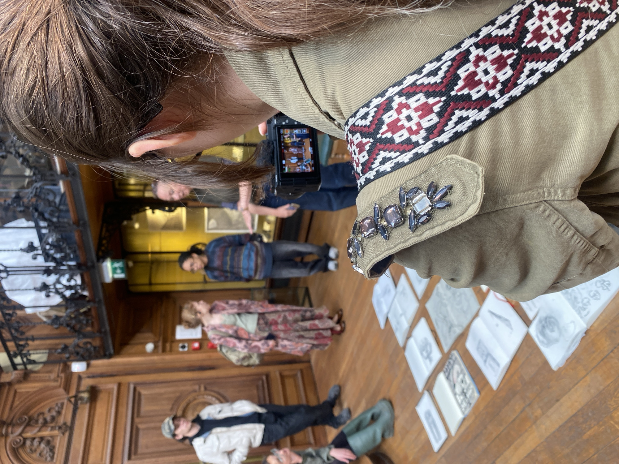 An overhead, slightly angled shot shows the back of a person's head and right shoulder, documenting an indoor scene with a digital camera. The person has long, brown hair and wears a khaki-colored jacket. The jacket features an ornate, woven strap with a geometric pattern in white, black, and maroon, running across the shoulder. The shoulder flap is embellished with several iridescent, possibly gemstone-like, beads in shades of gray and purple. The person is holding a compact digital camera, its screen displaying a miniature version of the scene in front of them.
The setting appears to be a grand room, possibly in a museum, gallery, or historical building, given the rich, dark wooden paneling on the walls and the polished wooden floor. To the left, an ornate dark metal railing or balustrade is partially visible.
Several other individuals are present in the midground and background, though they are somewhat out of focus. One person wears a striped sweater and jeans, another a long, floral-patterned dress, and a third a light jacket and cap. Scattered across the wooden floor in the foreground and midground are numerous pieces of paper, possibly sketches or small artworks, laid out for display or examination. The overall activity suggests people are observing and documenting an exhibition or a collection of creative works. No clear, readable text is visible on any signs or papers. The lighting suggests an indoor daytime setting.
