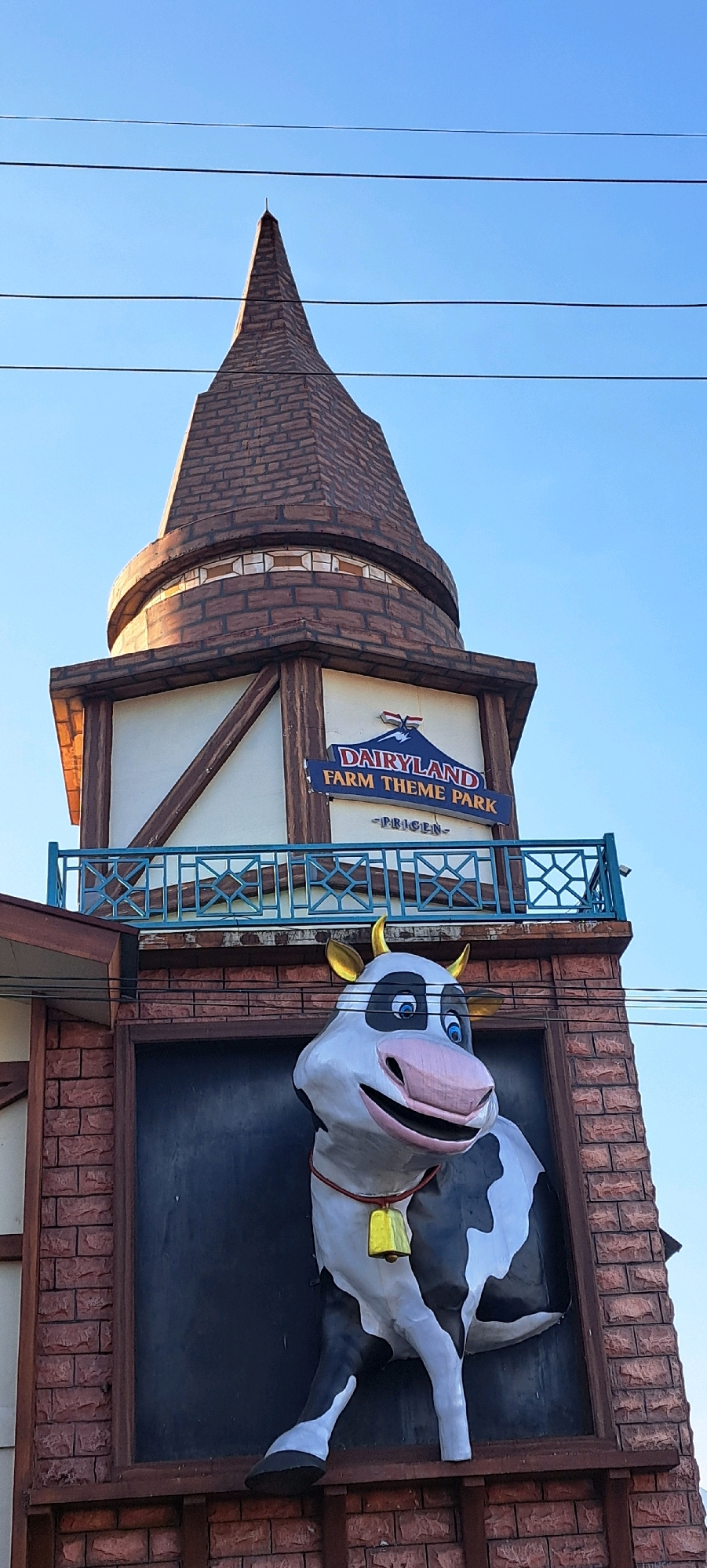 An exterior, low-angle, vertical medium shot captures the entrance facade of Dairyland Farm Theme Park in Prigen, Indonesia. The scene is dominated by a tall, pointed tower structure with a steeply pitched, dark brown roof resembling wood shingles, topped with a thin metal spire. The tower's midsection is white with dark brown timber framing, and a blue balcony with intricate geometric patterns runs across it. Below the balcony, a blue sign with white text reads "DAIRYLAND FARM THEME PARK - PRIGEN -".
At the base of the tower, a large, cartoonish cow statue is prominently displayed within a brick-like facade. The cow is black and white with large blue eyes, a pink snout, and gold horns. It wears a large yellow bell around its neck on a red string. The cow appears to be stepping forward out of a dark rectangular frame. The sky is a clear, pale blue with three dark power lines stretching horizontally across the top of the image. The overall impression is bright and cheerful, with a clear focus on the theme park's whimsical entrance.