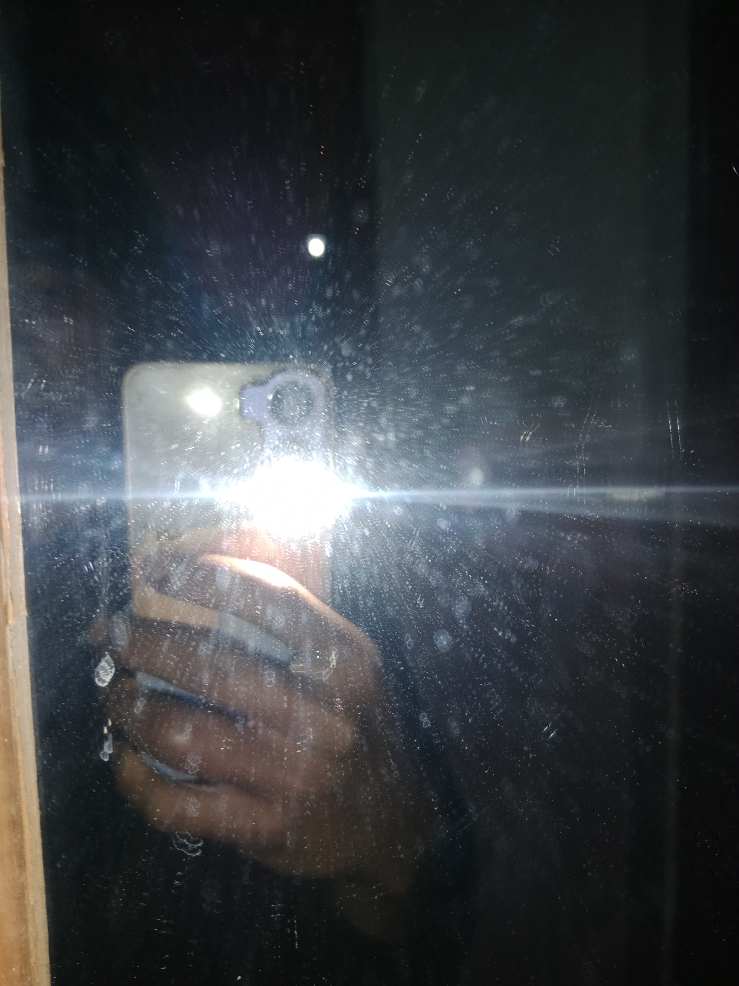 A reflection in a dark, dusty mirror shows a person holding a smartphone, taking a picture. The smartphone's flash is actively illuminating the scene, creating a bright glare and lens flare that obscures much of the detail. The person's hand, holding the phone, is visible. The mirror appears to be part of a doorway or furniture, with a wooden frame on the left side. The overall scene is dimly lit, with the primary light source being the phone's flash. The location is Ringinrejo, Indonesia.
