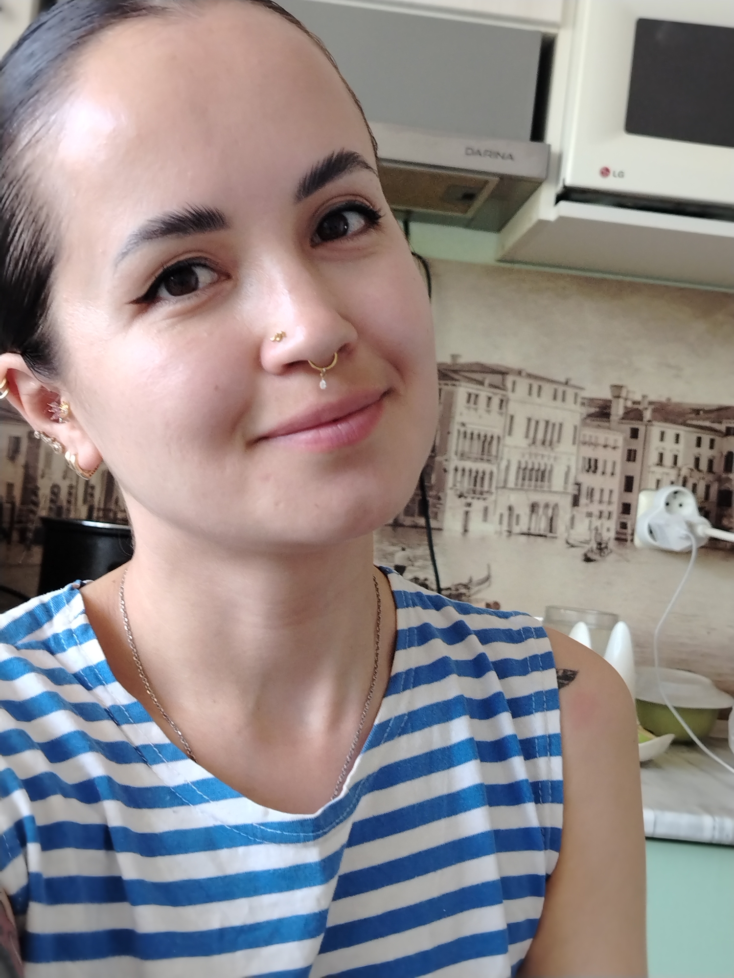 A close-up selfie depicts a woman with dark hair pulled back, wearing a blue and white striped tank top and a silver necklace. She has multiple piercings in her ears and nose, including a stud in her nostril and a ring in her septum. Her expression is a slight smile.
Behind her, a kitchen setting is visible. A wall covering features a printed cityscape of Venice, Italy. Kitchen appliances, including a range hood labeled "DARINA" and a white LG microwave, are positioned above a counter. A white electrical outlet and cord are also visible on the wall.