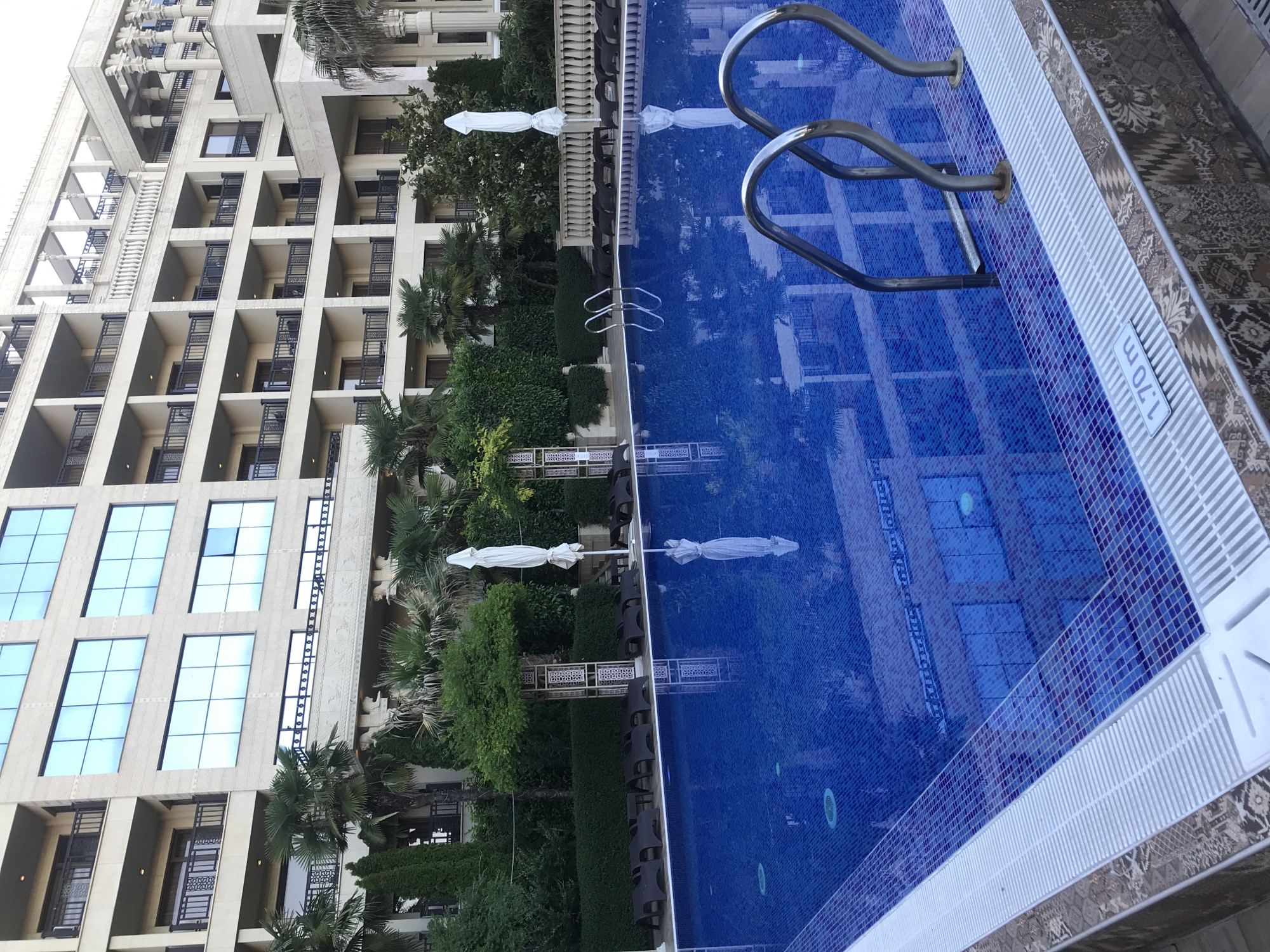 The image shows a swimming pool at the Grand Hotel Varna in Varna, Bulgaria. The pool is rectangular with blue tiled walls and a tiled border. There are stainless steel ladder handles extending into the water. Around the pool, there are several dark-colored lounge chairs arranged on the deck, and some white beach umbrellas are set up. In the background, a large, multi-story building with a light-colored facade and many windows is visible. The building appears to be a hotel or resort, with balconies on each level. Lush green trees and palm trees surround the pool and building, indicating a resort setting. A depth marker indicating "1.70 m" is visible on the pool's edge. The scene appears to be tranquil and well-maintained.
