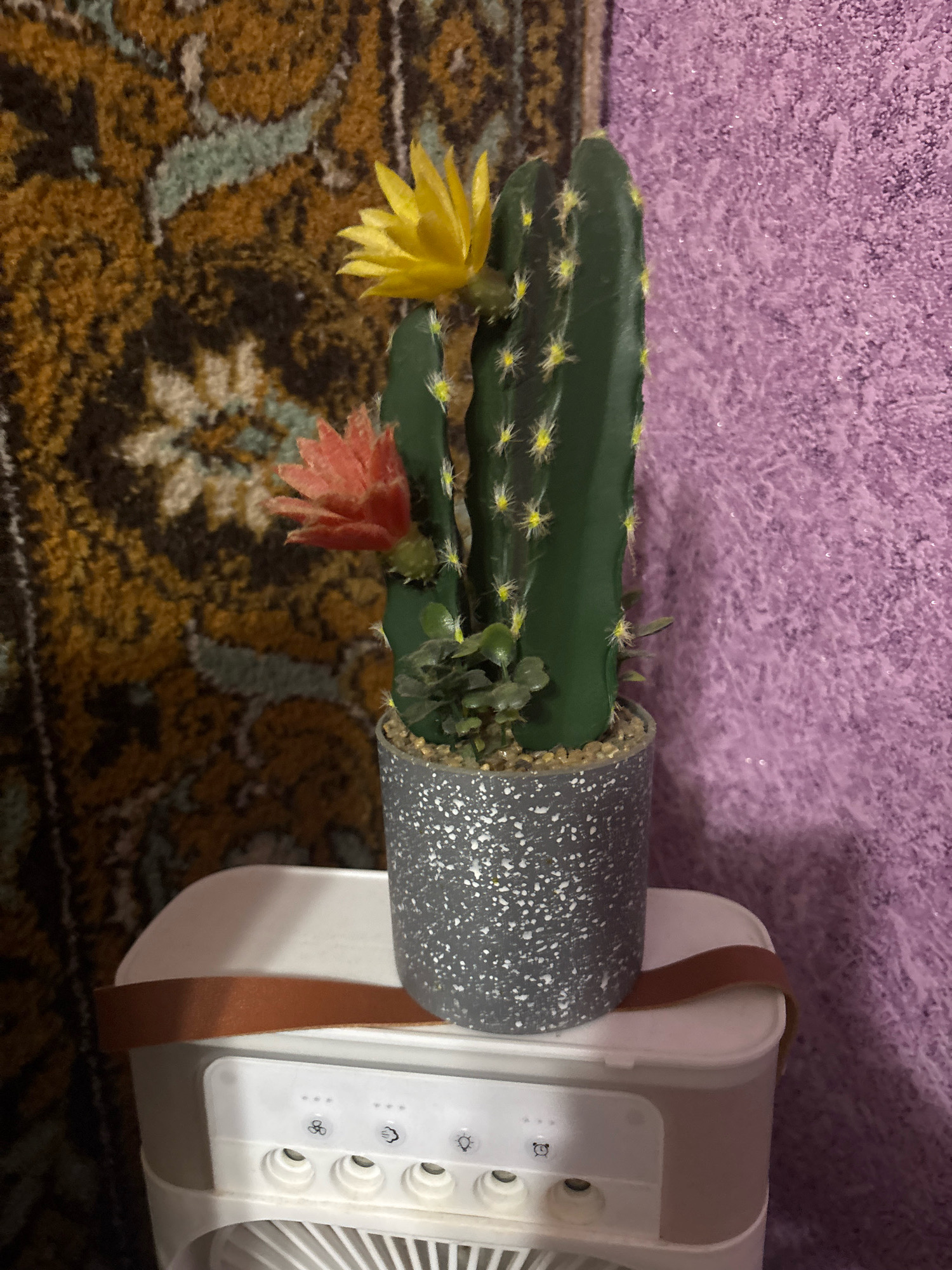 The image shows a close-up of a faux cactus plant in a speckled gray pot, placed on top of a white electronic device. The cactus has two artificial flowers: one yellow and one pink. The background consists of a textured purple surface on the right and a patterned rug with brown, gold, and green tones on the left. The white electronic device appears to be a small fan or air cooler with several buttons and indicator lights. A brown strap is visible around the top of the device, securing it. The scene is indoors, with the lighting suggesting it was taken with flash. There is no identifiable information about the location in Novocheboksarsk, Russia.