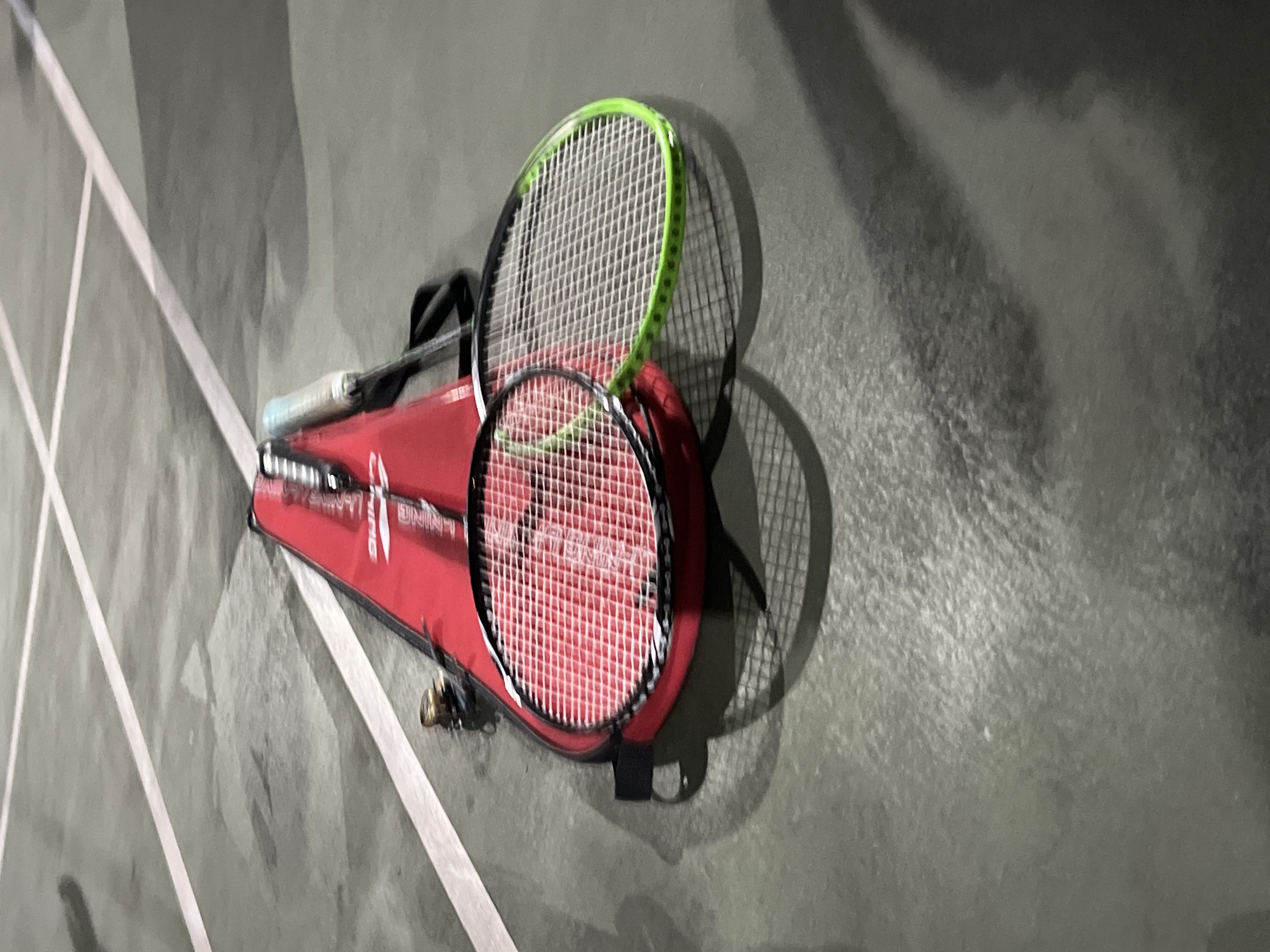 The image shows two badminton rackets resting on a red badminton bag. The bag and rackets are on a gray, outdoor court, likely a badminton court, with white lines visible on the surface. The scene is outdoors, and the lighting suggests it might be evening or late afternoon. There are no people visible in the image. The overall scene is static, depicting sports equipment on a court. The Yuzhong District is a district of Chongqing, China.