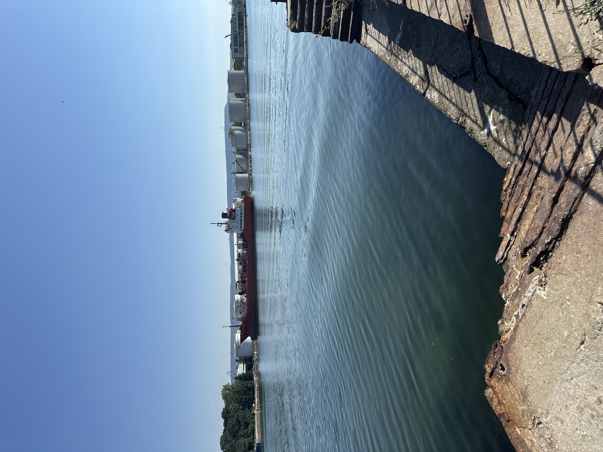 This is a full, eye-level, outdoor shot of a calm harbor on a clear, sunny day. The water in the foreground is a deep blue-green with subtle ripples. In the middle ground, a long, red cargo ship is docked, its bow facing left and stern to the right. Behind the ship, a line of large, white cylindrical storage tanks is visible on the shore, suggesting an industrial or port area. Further in the distance, a line of green trees and hills forms the horizon under a clear, bright blue sky. The right side of the foreground shows a concrete dock with weathered steps leading down to the water, and some rusty metal structures are visible on the edge of the dock. The overall impression is one of industrial activity in a serene waterfront setting.