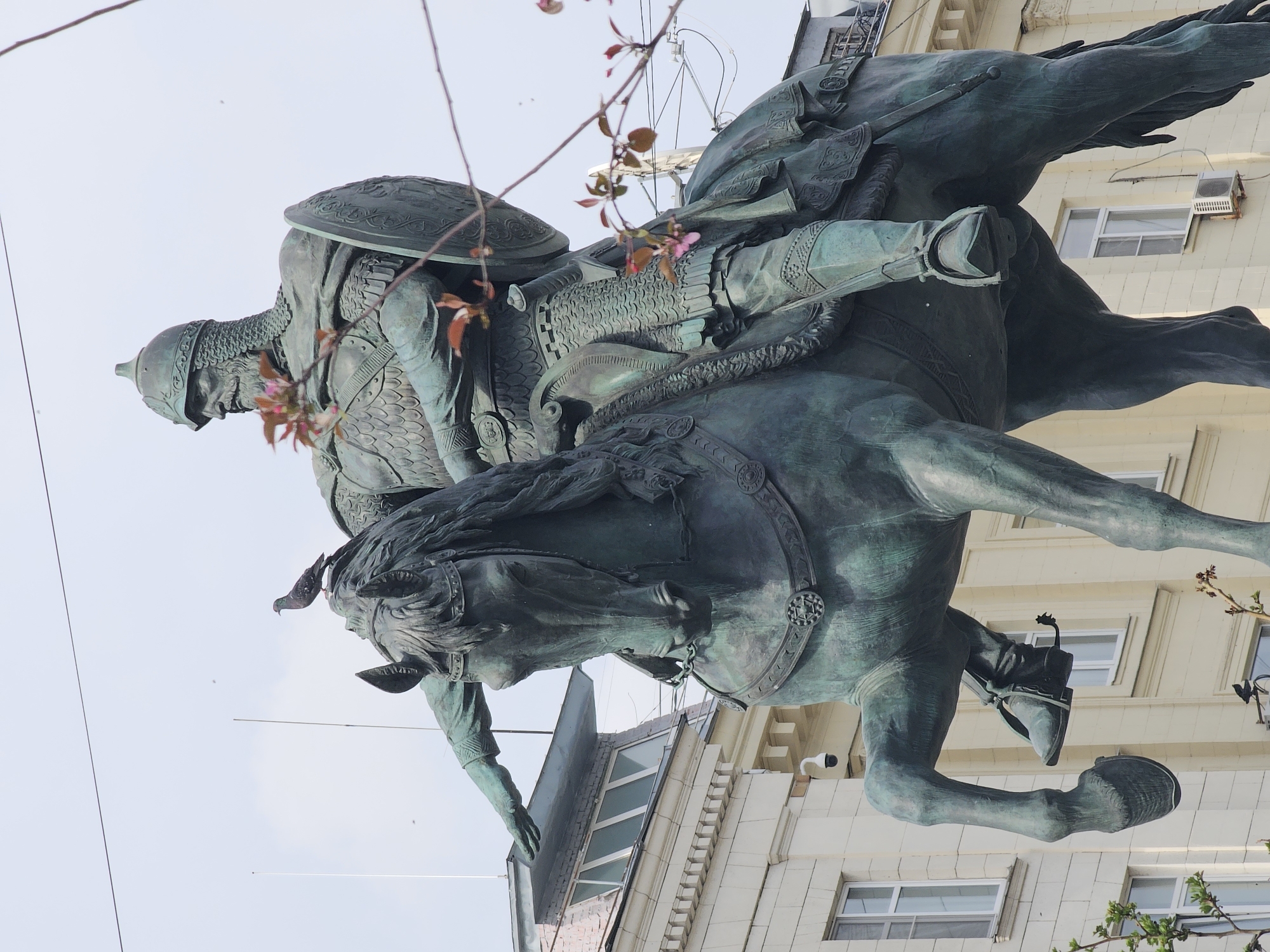 The image shows a statue of Yuri Dolgoruky on horseback in Moscow, Russia. The statue depicts a medieval warrior in full armor, including a helmet, shield, and chainmail. He is seated on a horse, which is rearing up on its hind legs. The statue is made of bronze and has a greenish patina. In the background is a building with white walls and windows. Branches with pink blossoms are in the foreground, obscuring parts of the statue. A bird perches on the head of the horse. Power lines are also visible against the sky. The statue appears to be facing to the left of the frame.