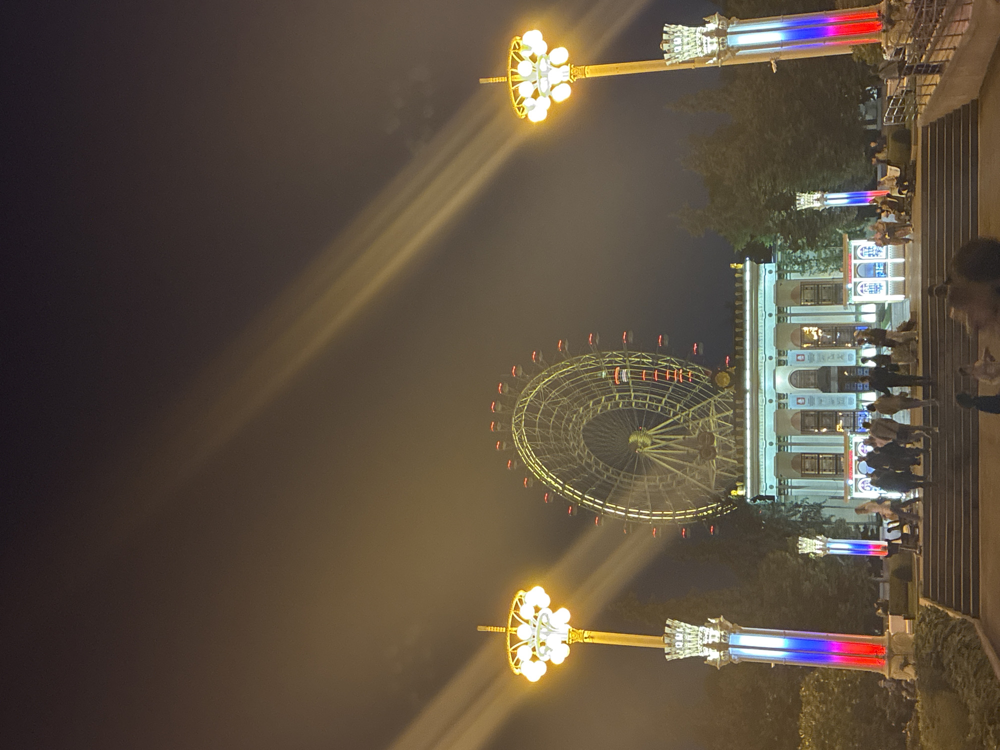 This nighttime photograph captures the iconic Ferris wheel at VDNKh (Exhibition of Achievements of National Economy) in Moscow, Russia. The Ferris wheel, illuminated with red and yellow lights, dominates the background, its large circular structure a prominent feature against the dark sky. Beams of light, likely from spotlights or projectors, cut diagonally across the hazy atmosphere, adding a dramatic effect to the scene.
In the foreground, a grand white building with classical architectural elements serves as a focal point. People are gathered on a wide staircase leading up to this building, some walking and others standing in groups. To the left and right of the staircase are tall, ornate lamp posts topped with clusters of bright yellow lights. These lamp posts are also decorated with vertical stripes of red, white, and blue lights, resembling the Russian flag. Lush green trees frame the scene on either side, their dark silhouettes contrasting with the illuminated structures. The overall atmosphere is one of a vibrant, illuminated public space at night, likely a popular destination for recreation and sightseeing.