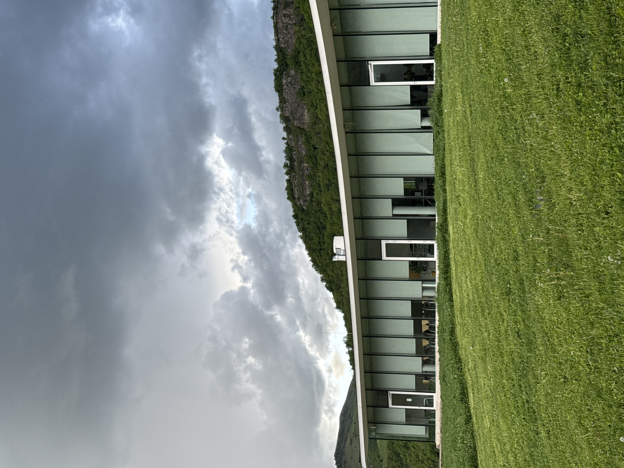 The image shows a building with a curved facade and large windows, adjacent to a grassy area. Behind the building, there is a cliff face with green vegetation. The sky is filled with dark, dramatic clouds. The building's interior appears to have tables and chairs. The scene is most likely located in Chkalov, Armenia, given the prompt.