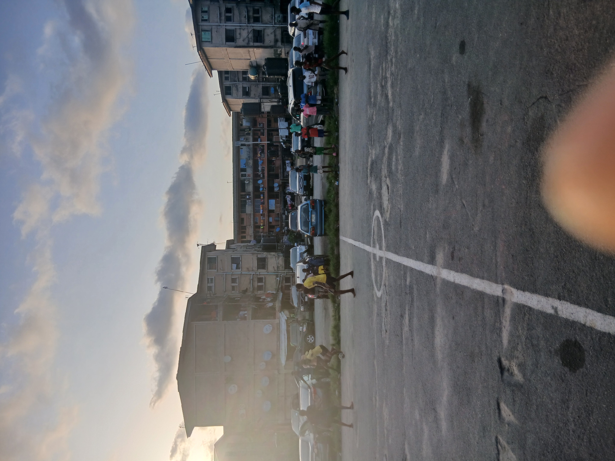 The image captures an outdoor urban scene, likely in Lagos, Nigeria, during late afternoon or early evening, indicated by the soft, warm light and a clear sky with scattered clouds.
In the foreground, a paved surface, possibly a basketball or general sports court, features visible white lines including a straight boundary and part of a circular marking. Several individuals, appearing to be young men or boys, are present. Two figures in the mid-ground are central to the activity, one prominently wearing a yellow shirt, suggesting they are engaged in a casual game or physical activity on the court. Other people are standing or walking along the edge of the court.
The background is dominated by multi-story residential buildings, some appearing aged, with multiple windows, balconies, and possibly laundry hanging outside. A row of parked cars is visible along the side of the court, bordering the buildings. The overall setting depicts a busy, communal urban environment.
No discernible text is visible on any objects, clothing, or signs within the image. A blurred human finger or thumb partially obscures the lower right corner of the frame.