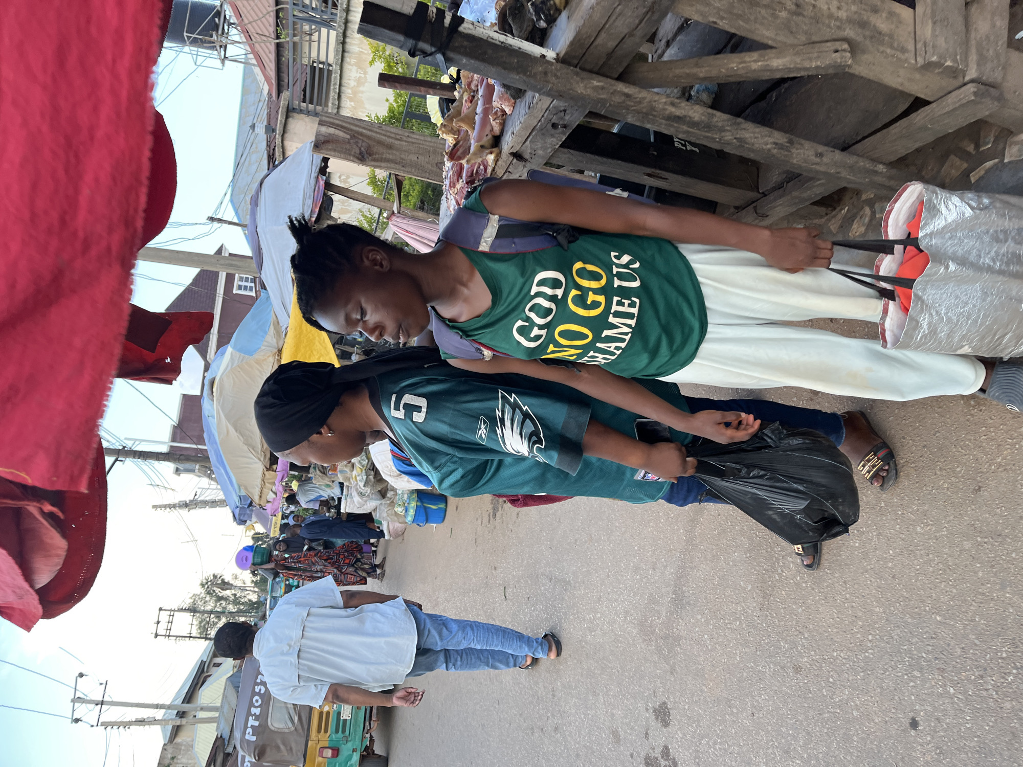 The image depicts a street scene in the city of Jos, Nigeria. In the foreground, two young women stand on a paved street. The woman on the left wears a dark headscarf and a green Eagles jersey with the number 5. She is holding a black plastic bag in her left hand and her right hand is visible near her waist. The woman on the right wears a green sleeveless shirt with white and yellow lettering that reads "GOD NO GO SHAME US." She has a backpack on and is holding a silver shopping bag with red items inside. Behind them, a man in a white shirt and blue jeans walks away from the camera. In the background, a colorful auto-rickshaw with the license plate "PT1057" is visible, along with market stalls displaying raw meat. Various umbrellas and overhead coverings suggest a bustling marketplace environment. The sky is visible with some clouds.