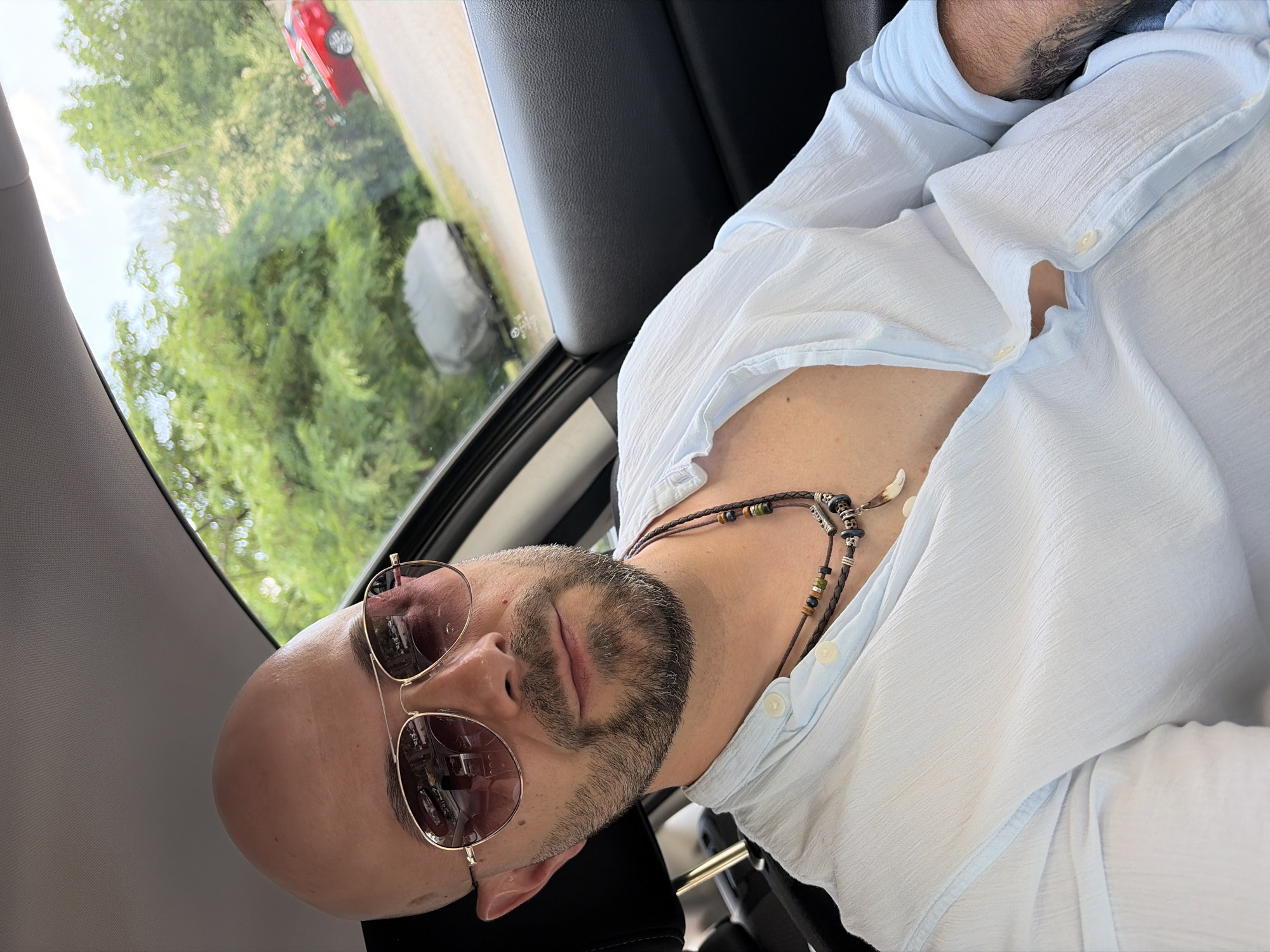 A man with a shaved head, sunglasses, and a goatee is sitting in a car. He is wearing a light blue, short-sleeved button-down shirt and a beaded necklace. The car appears to be in motion, as indicated by the blurred trees and a red car visible through the window. The lighting suggests it is daytime. The man's eyes are closed, giving him a relaxed or contemplative appearance. The interior of the car is visible, including a black leather seat and a portion of the dashboard. The view through the window shows lush green foliage, implying a rural or suburban setting. The weather appears to be clear.