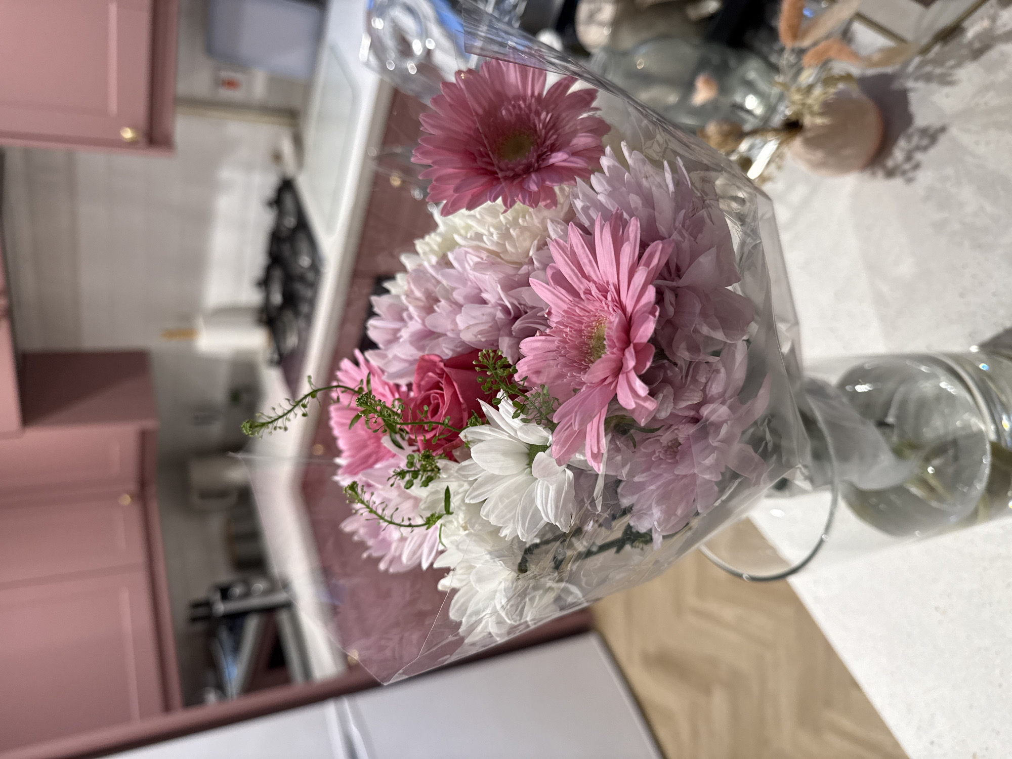 The image depicts a bouquet of flowers in a clear vase, situated on a light-colored countertop. The bouquet includes pink and white chrysanthemums, a pink gerbera daisy, and red roses, wrapped in clear cellophane. The countertop surface appears to be light grey or white with subtle variations in tone, possibly representing a textured or patterned material. Background details indicate a domestic kitchen setting, with pink cabinets visible on the left and glimpses of kitchen appliances (likely a stove and refrigerator) in the background. The flooring in the background is visible as light-colored wooden planks. The image does not depict any people, actions or interactions. The location is not explicitly confirmed but is hypothesized as East Suffolk, United Kingdom based on background details consistent with typical domestic interiors and kitchen design.