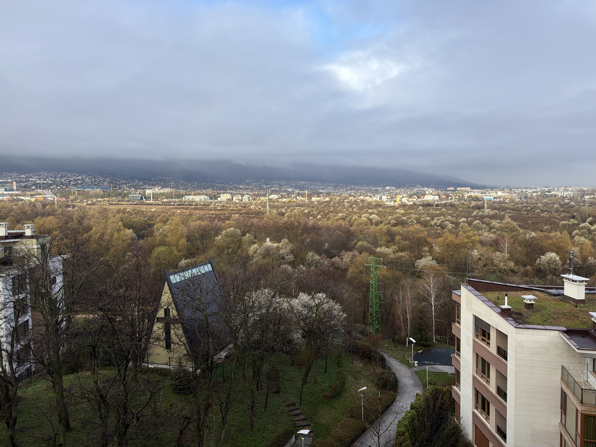 The image shows a cityscape in Sofia, Bulgaria, under a cloudy sky. In the foreground are several buildings and trees. A church with a prominent cross is visible among the trees. A winding paved pathway runs through the landscape. In the distance, the city of Sofia stretches towards the base of a mountain range partially obscured by clouds. An electricity pylon can be seen in the middle distance. The city's buildings are densely packed, gradually fading into the cloud cover. The trees in the middle ground appear to be in early spring, with some showing blossoms. The architecture includes residential buildings with various designs. The roofs of some buildings in the immediate foreground are covered with vegetation. There are no people visible in the image.